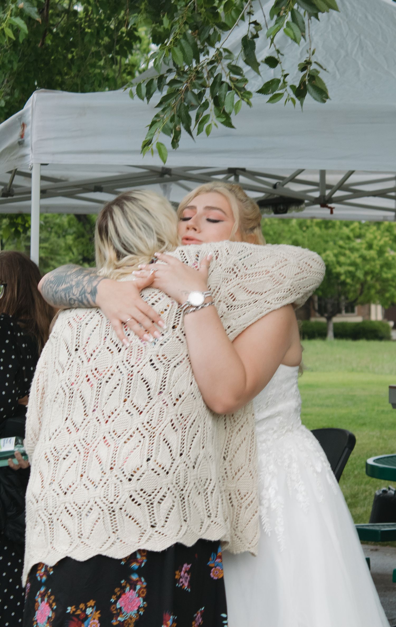 A heartfelt hug shared between wedding attendees wearing a white dress and knit shawl under an outdoor canopy.