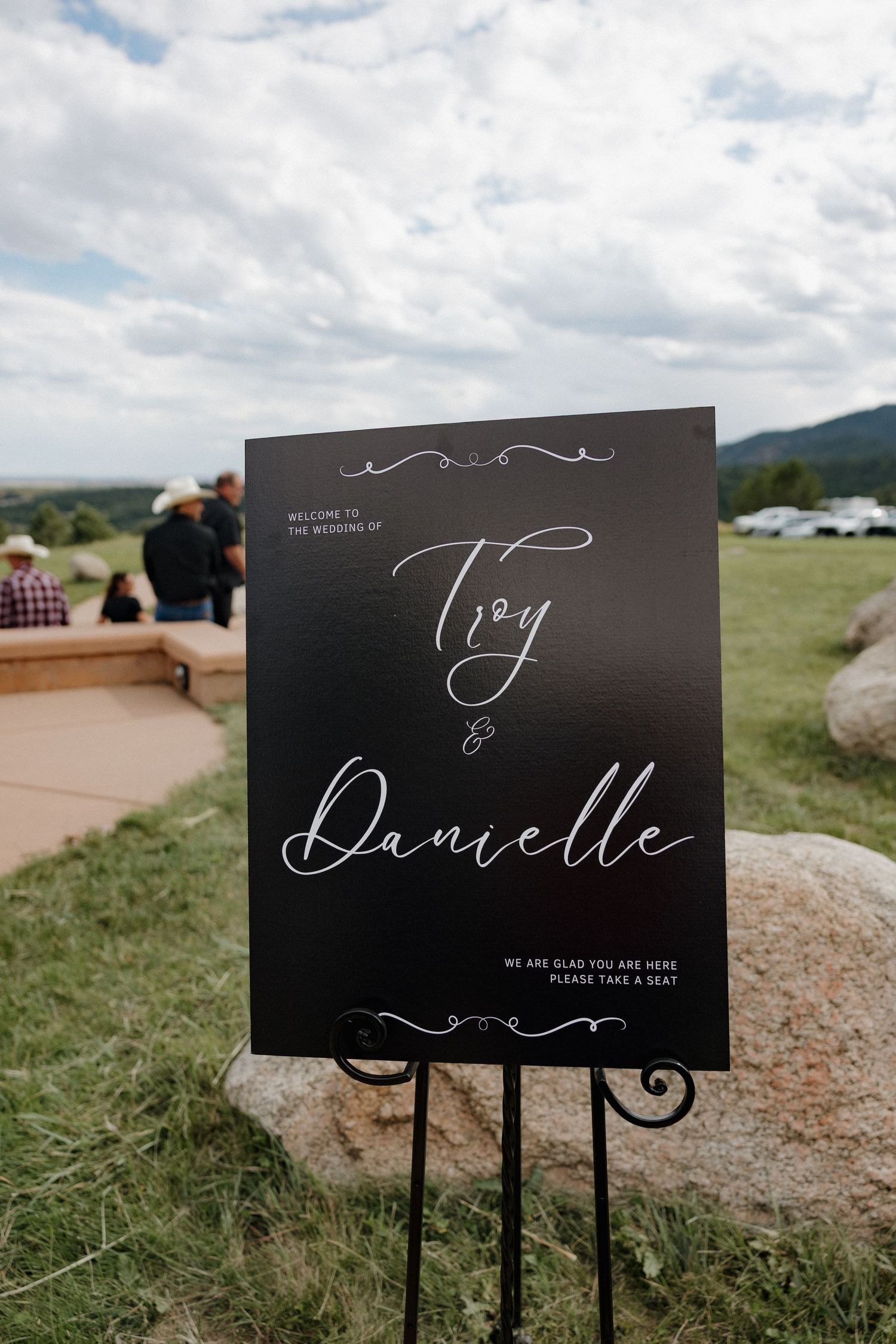 Elegant black wedding welcome sign for Troy and Danielle displayed on an easel at an outdoor ceremony.