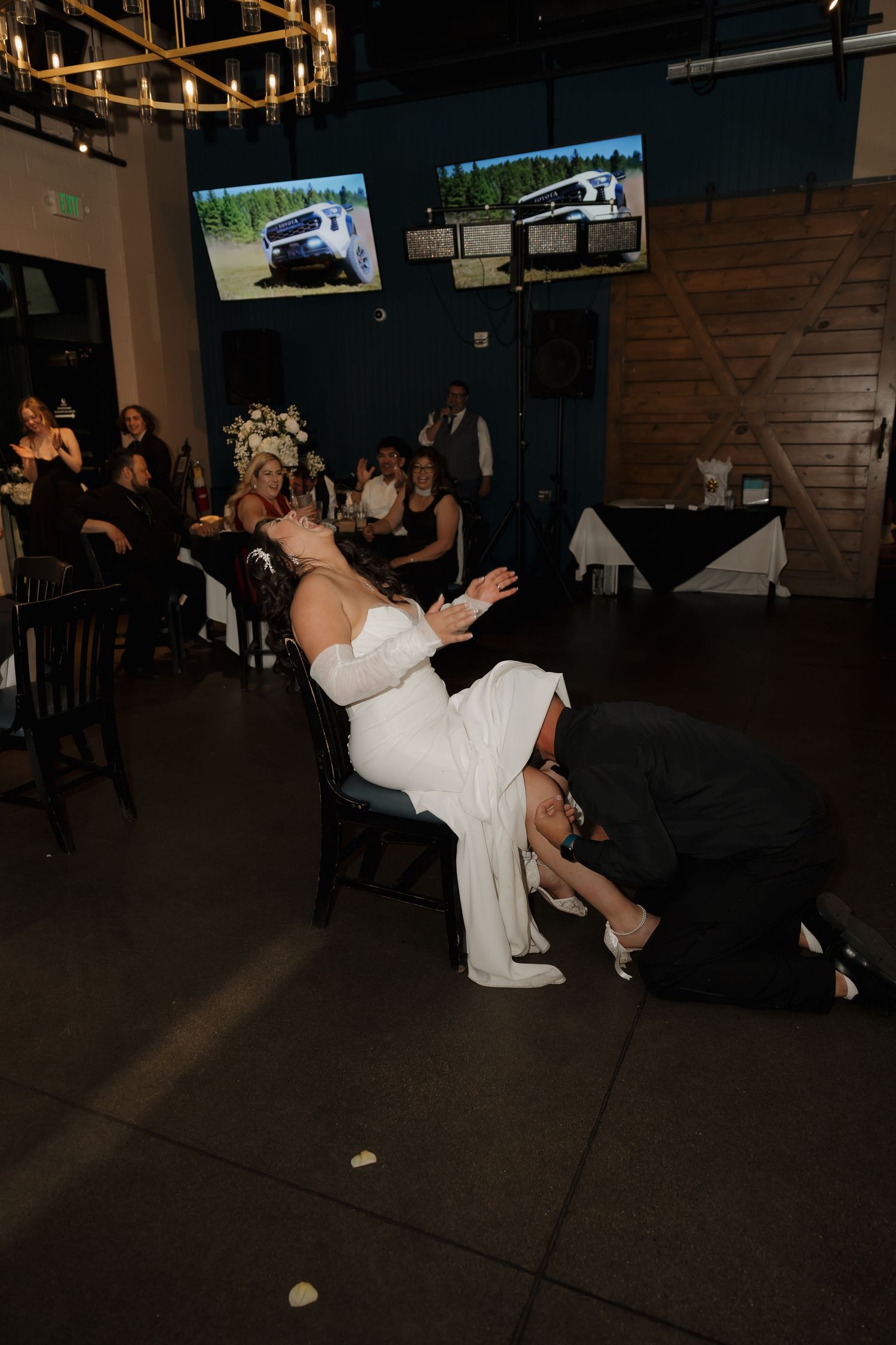 Groom kneels to remove garter from bride seated in chair during fun wedding reception tradition.