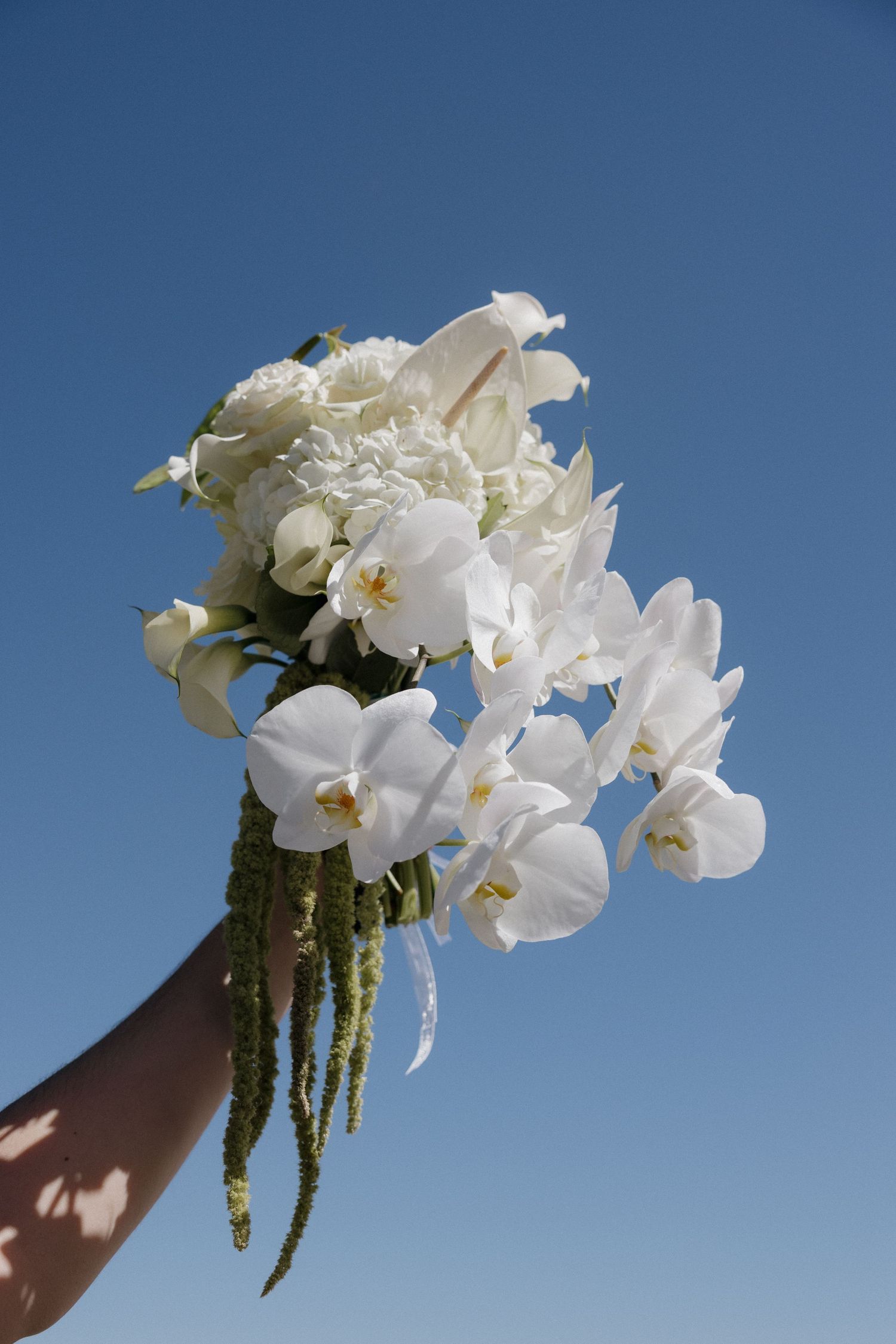 Hand holding a bridal bouquet of white orchids and roses against a clear blue sky.