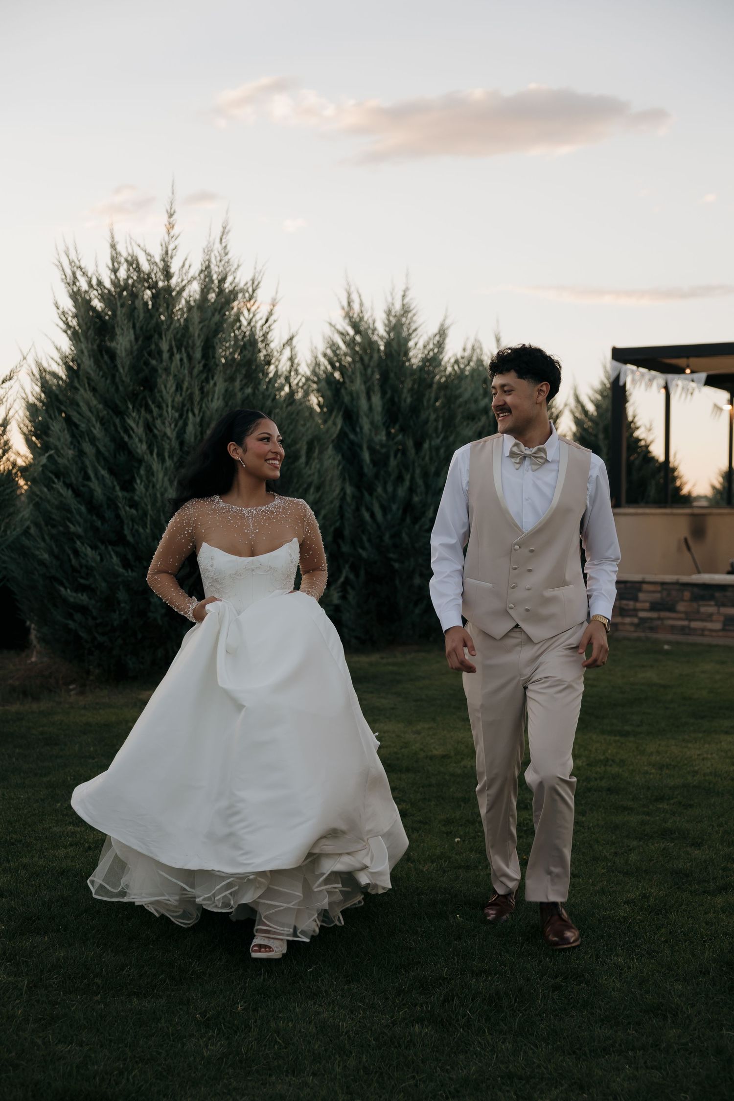 Bride and groom walk together laughing at sunset with evergreen trees in the background.