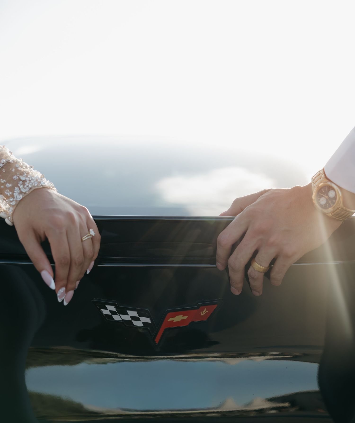 Bride and groom's hands rest on black Corvette hood, showcasing wedding rings in sunlight.