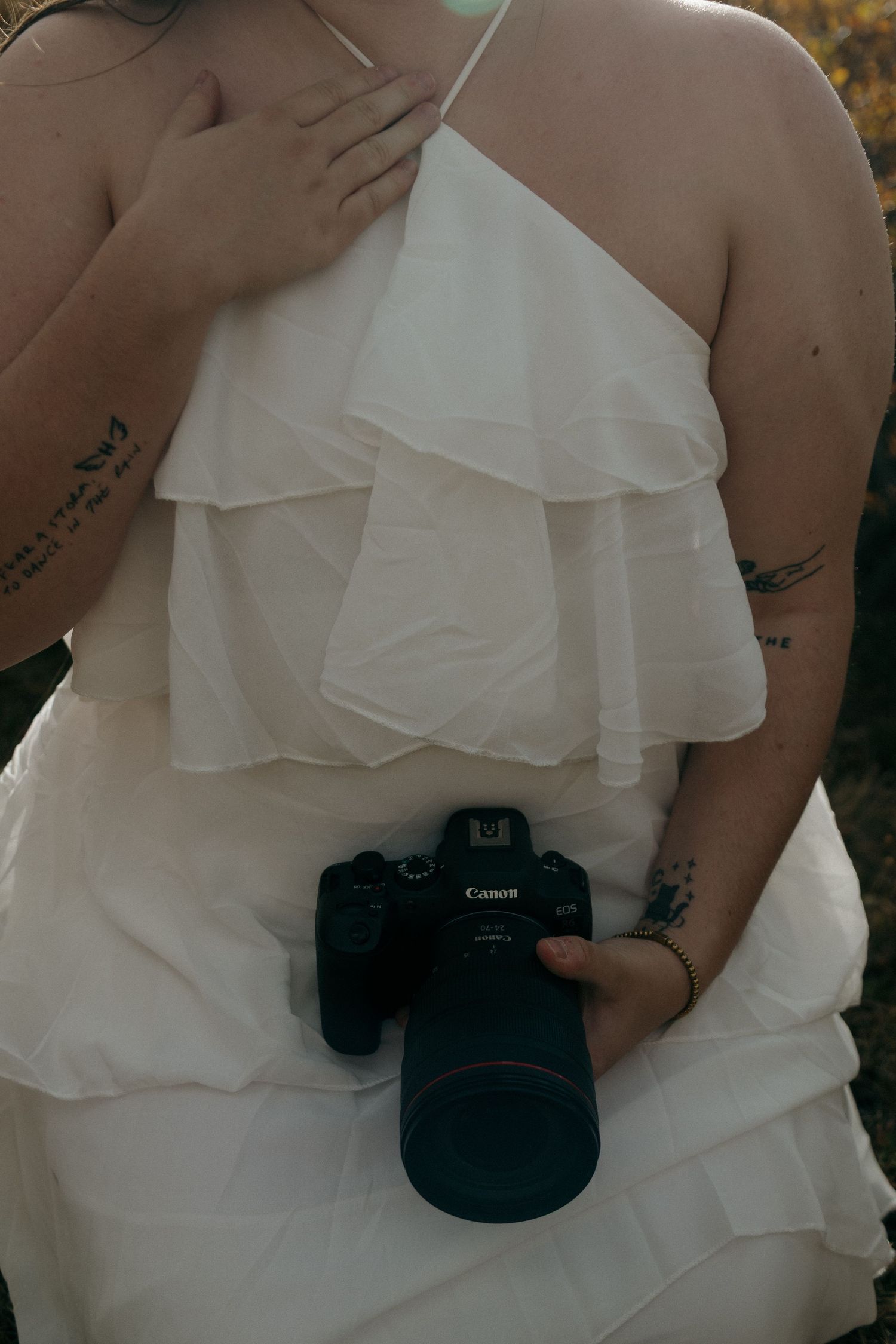 Woman in white ruffled halter dress holding a Canon DSLR camera, with visible tattoos on her arms.