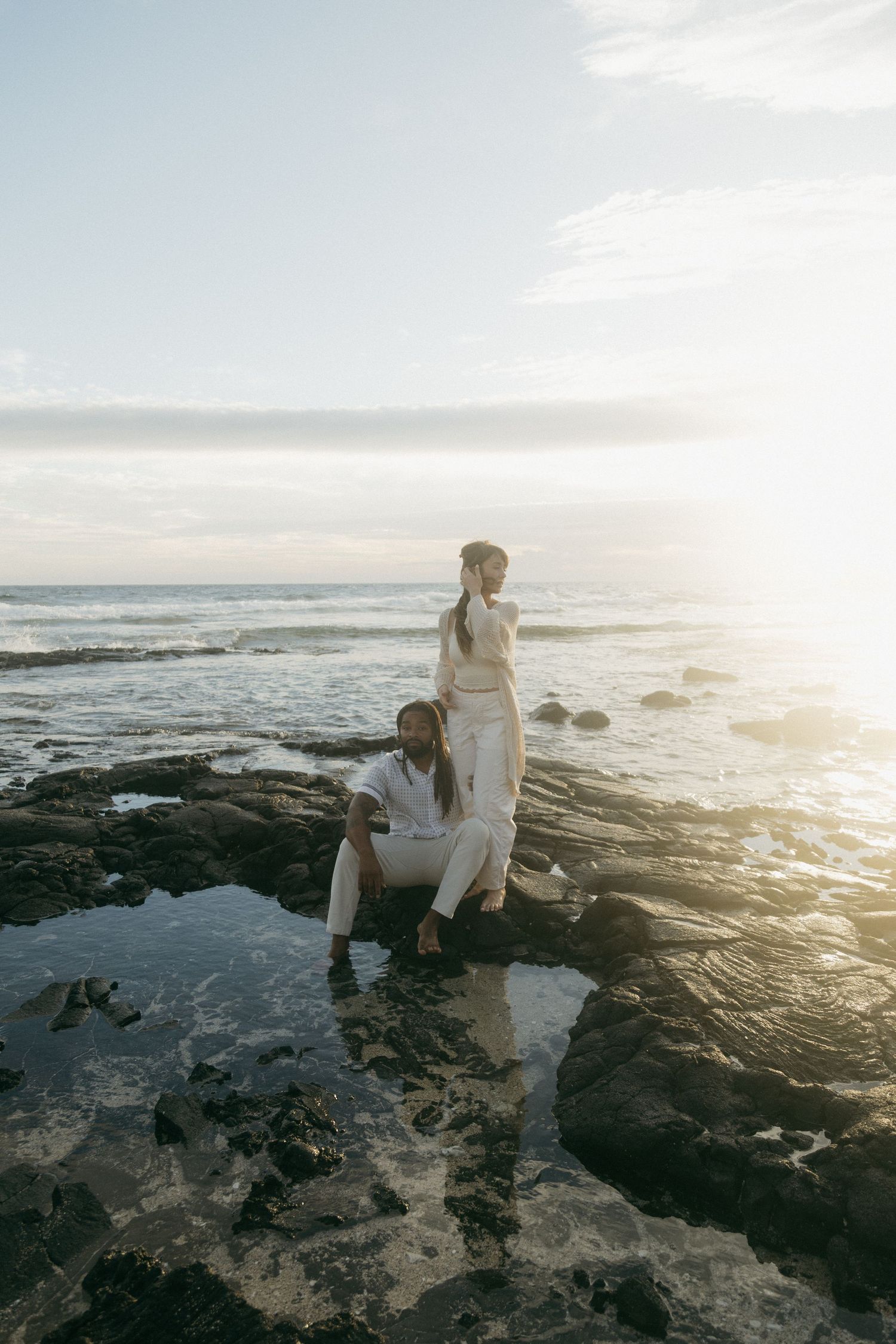 People sitting on rocky coastal shoreline at sunset with waves crashing against dark volcanic rocks in Hawaii.