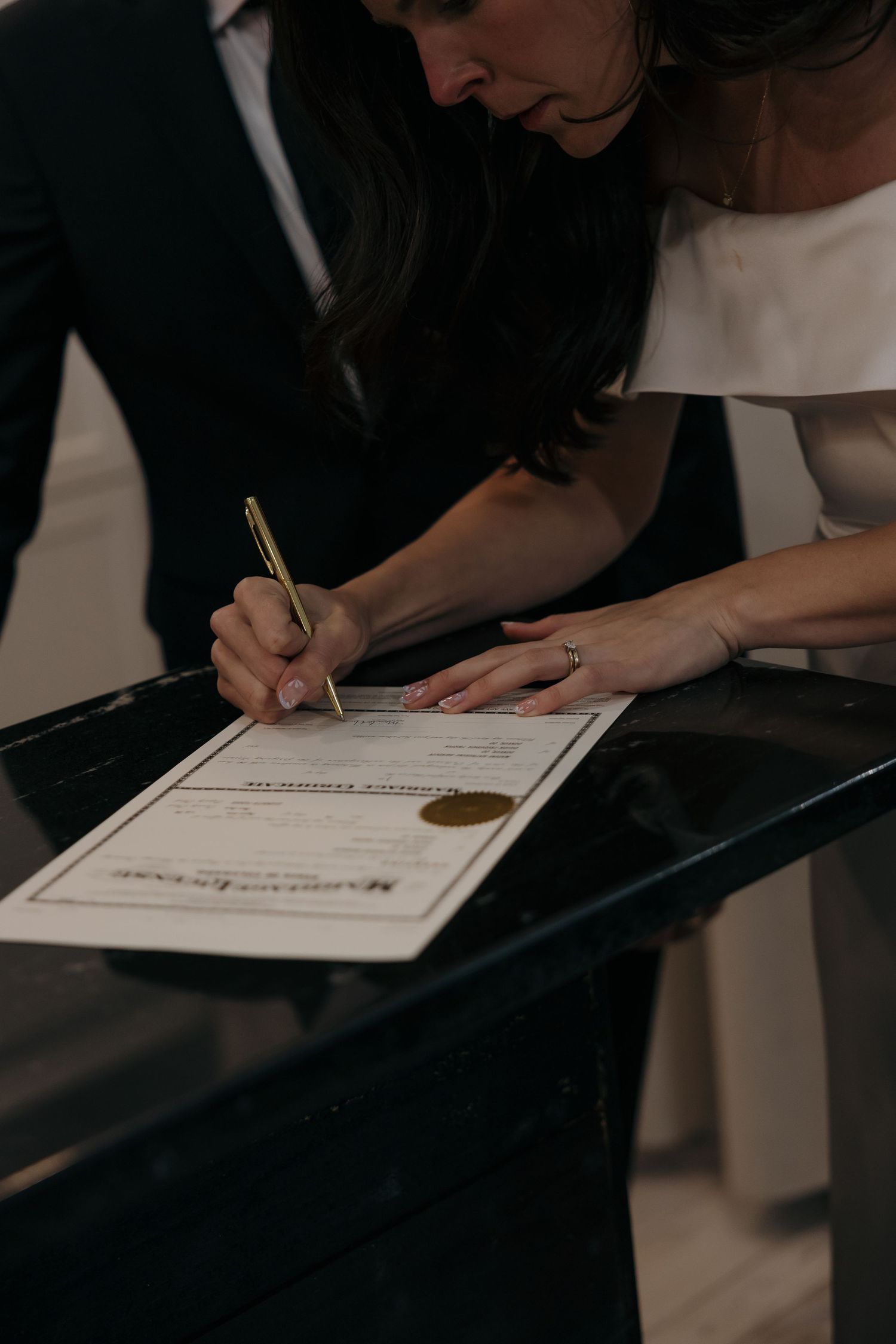 Bride signing marriage certificate with gold pen at elegant dark table during wedding ceremony.