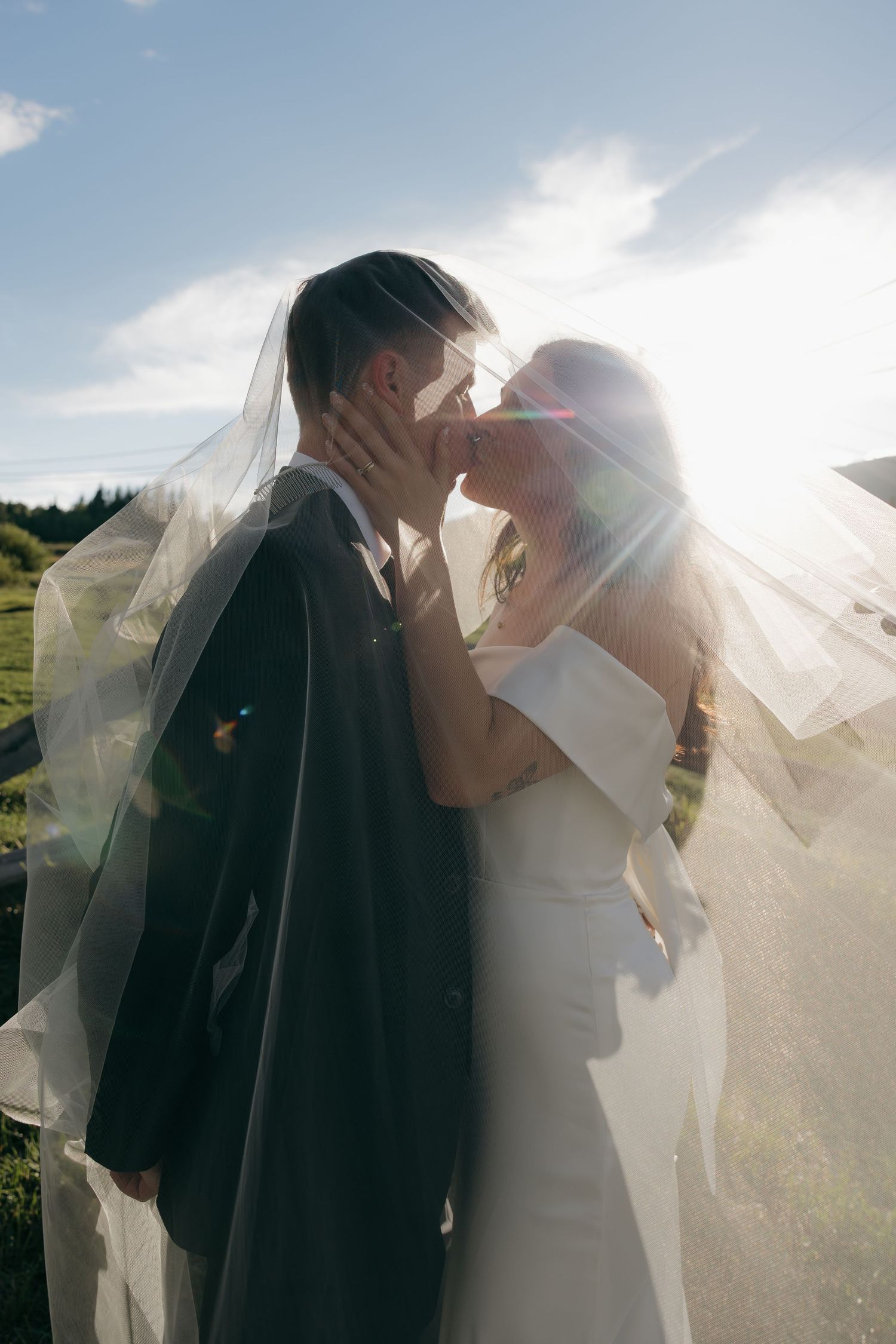 Wedding couple kissing under flowing veil with sunlight streaming behind them in outdoor ceremony.