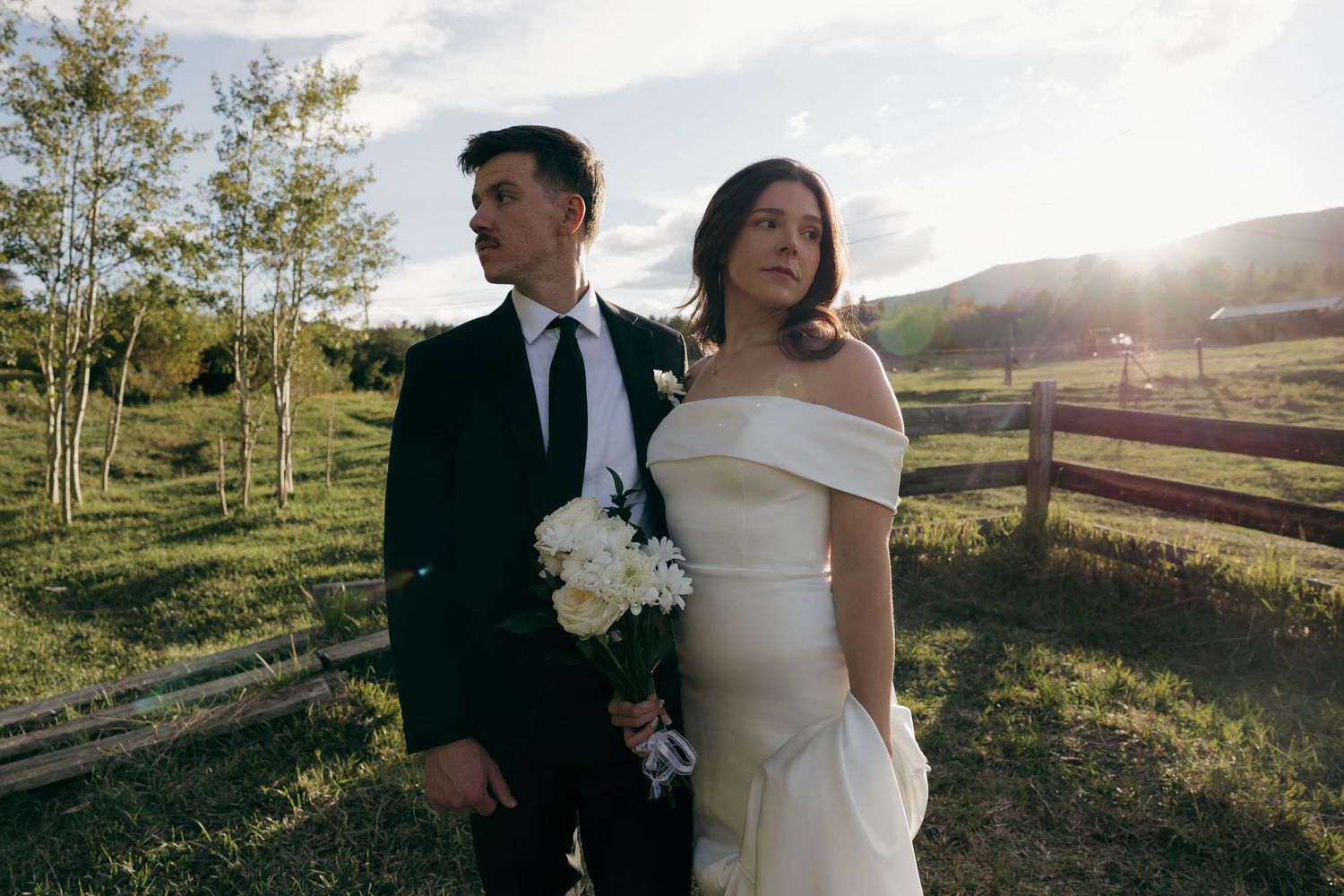 Bride in off-shoulder gown and groom in black suit pose at golden hour near fence.