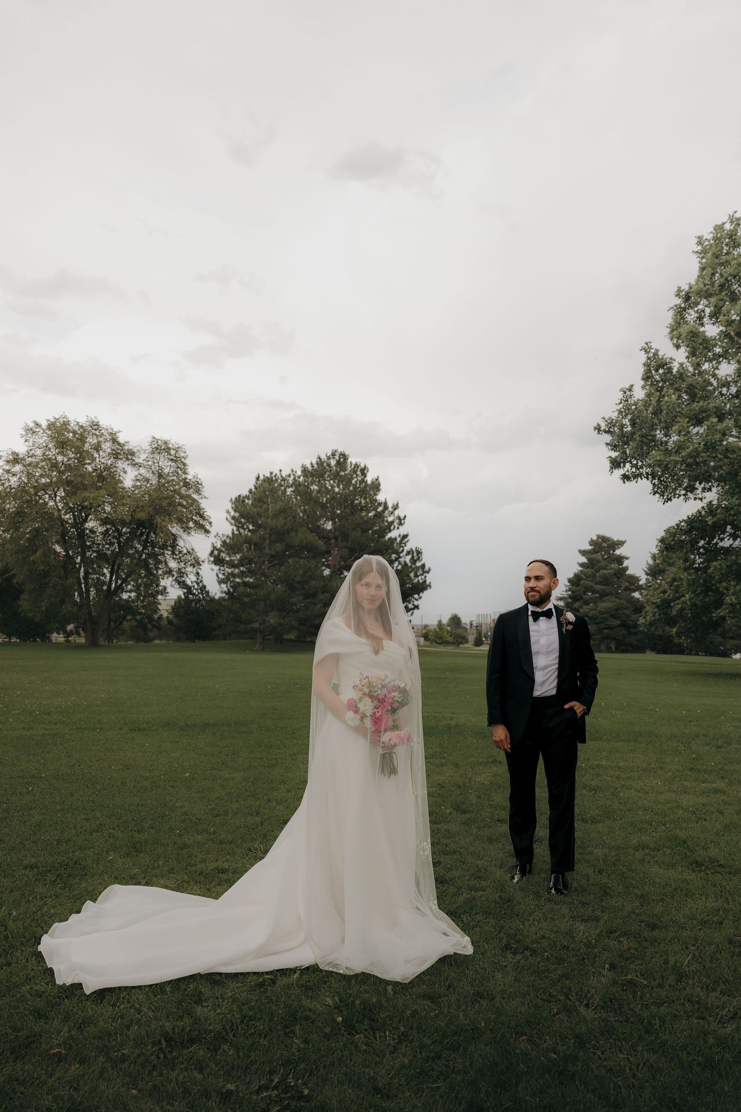 Bride in cathedral veil holding pink bouquet stands with tuxedo-clad groom on lush green lawn.
