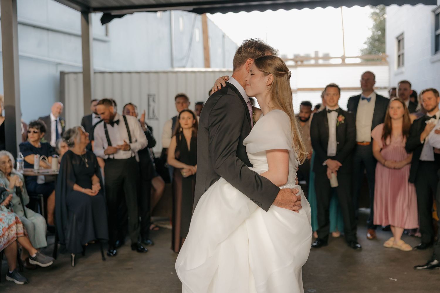 Bride and groom share their first dance surrounded by guests in a modern industrial wedding venue.