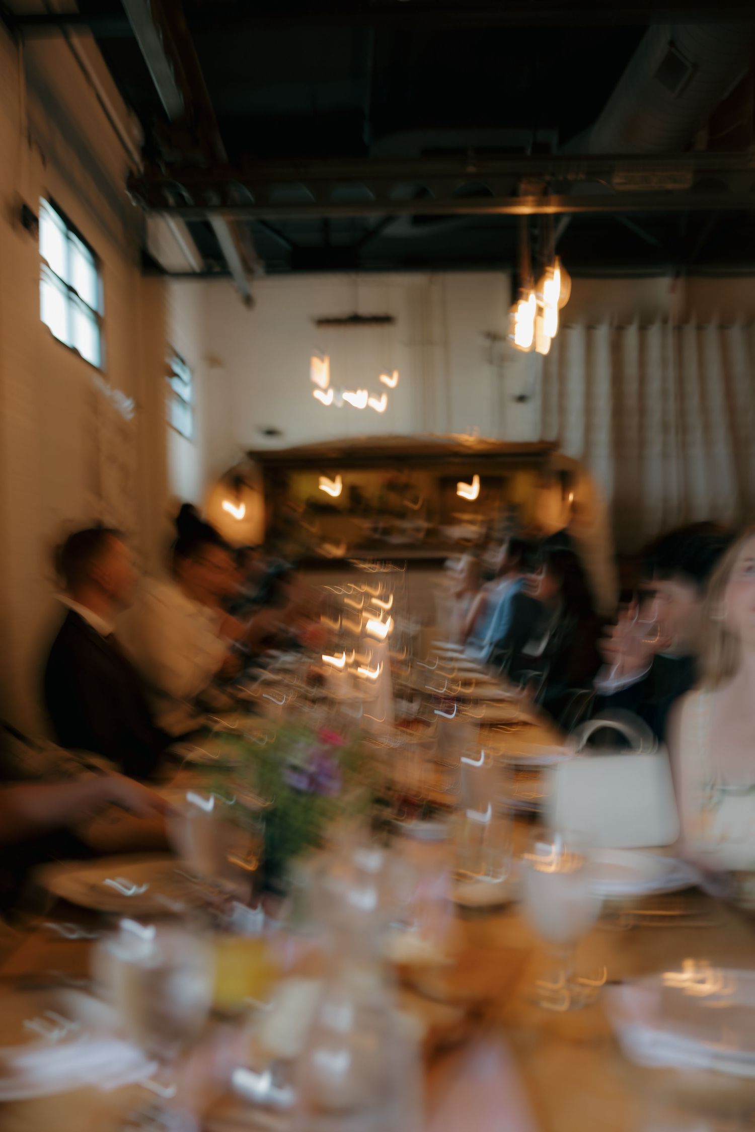 Blurred artistic shot of wedding reception guests seated at candlelit dinner table in warmly lit venue.