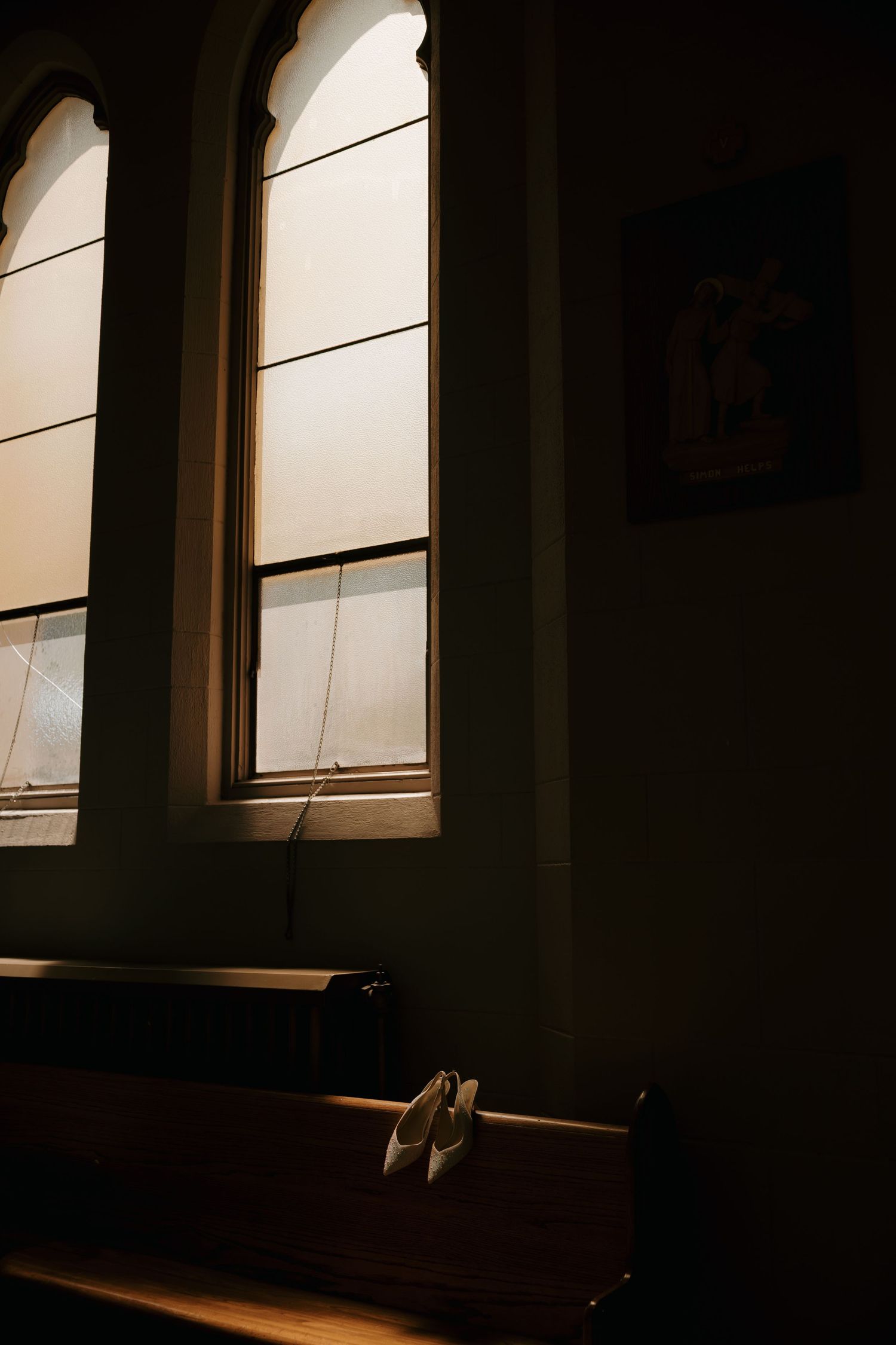 White bridal shoes resting on a wooden church pew beside gothic arched windows in a dimly lit chapel.