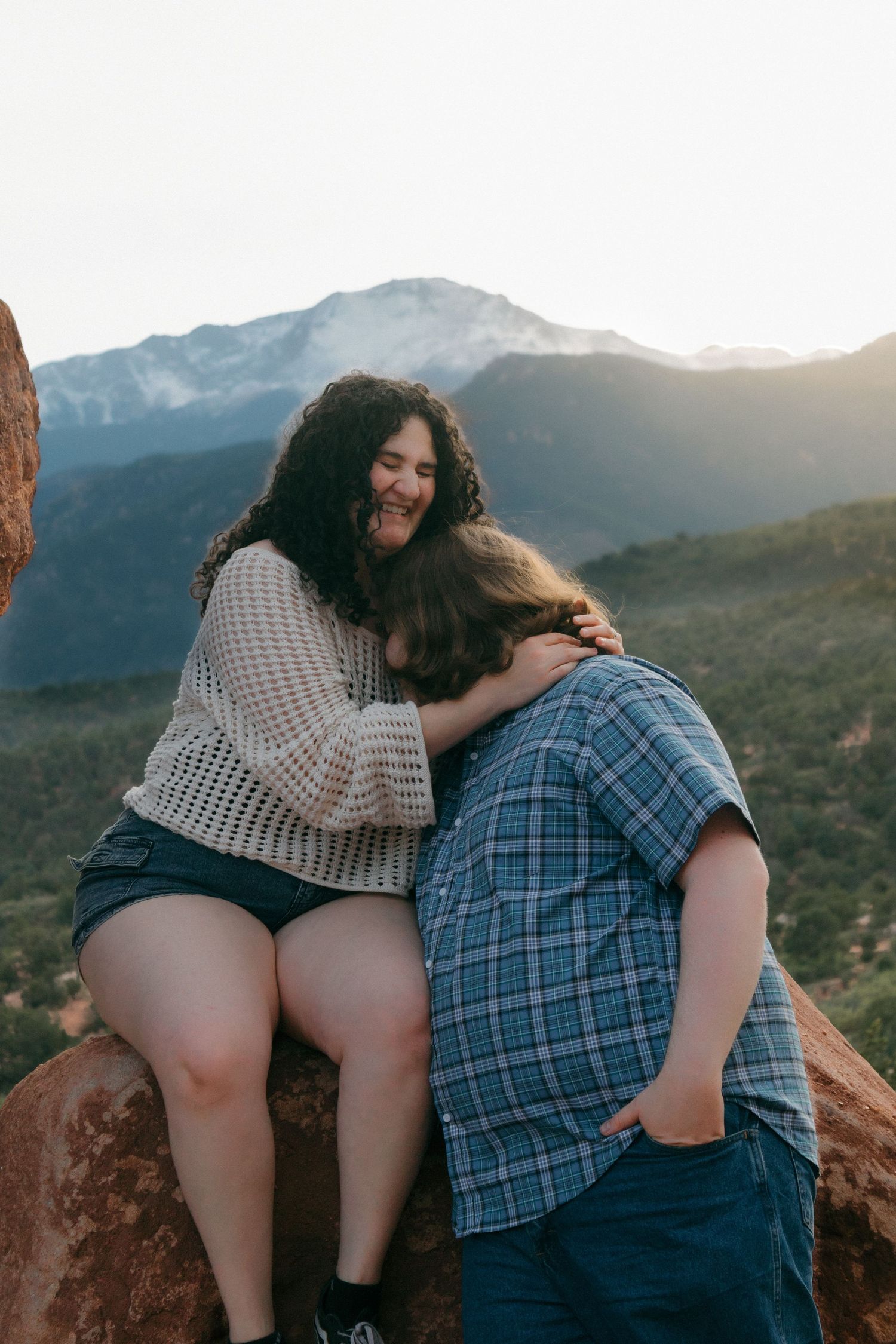 A playful couple shares a joyful moment while sitting on red rocks with mountains in the background.