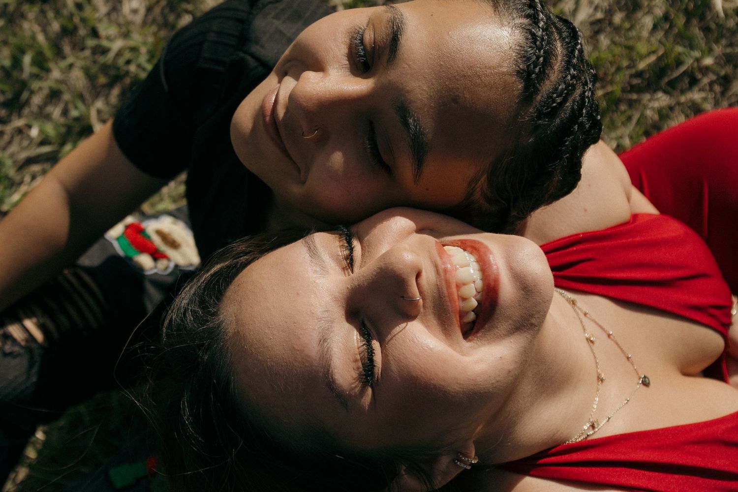 Two smiling women lying on grass together, sharing a joyful moment outdoors in natural sunlight.