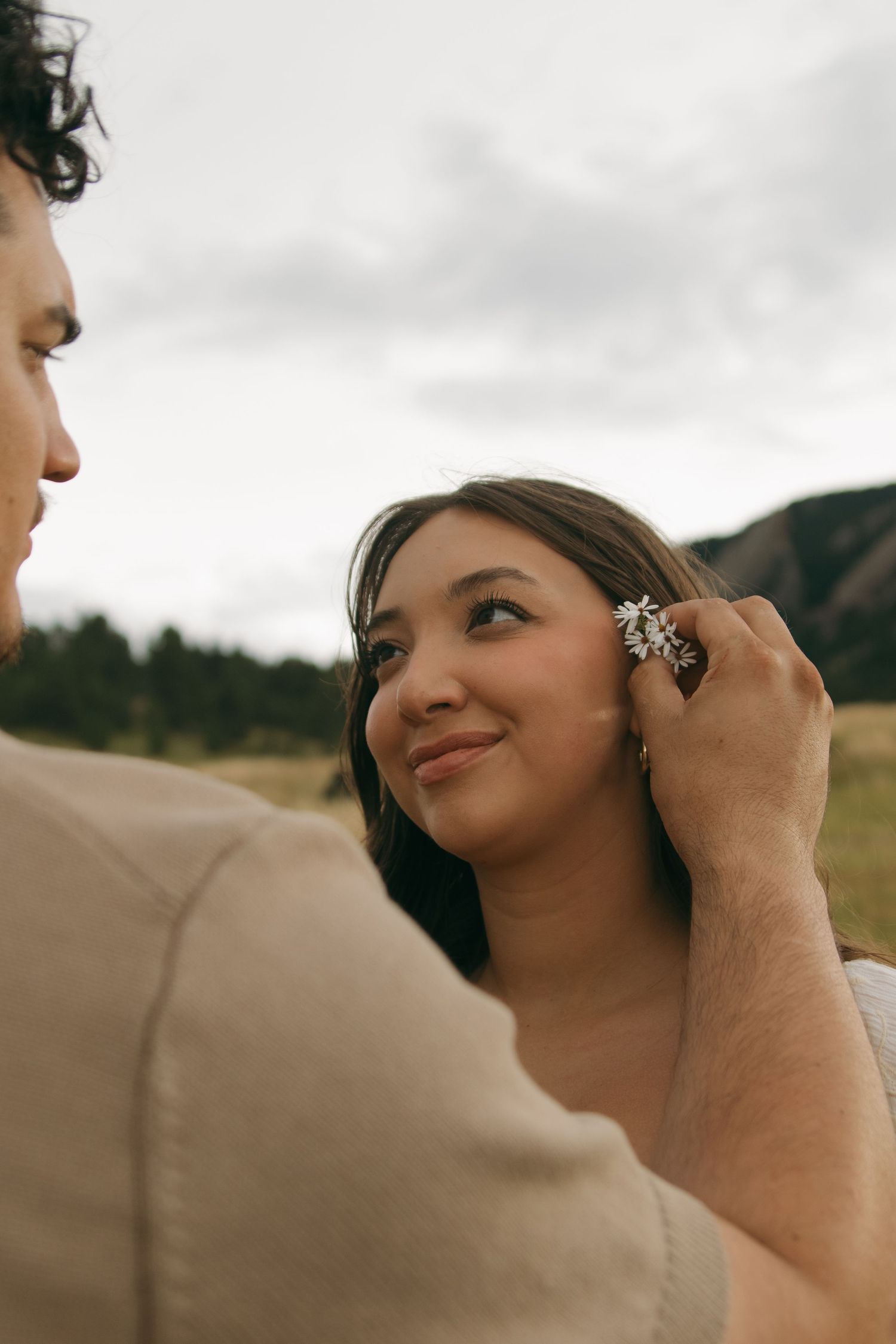 Man tucking a small white flower behind a smiling woman's ear in a mountain field.