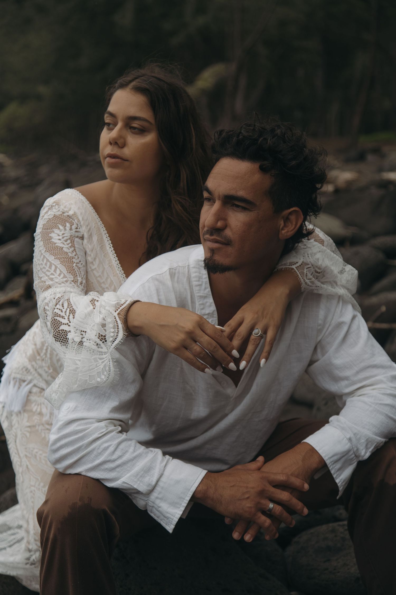 Bride in lace wedding dress embracing groom from behind while sitting on rocks in a forested setting.