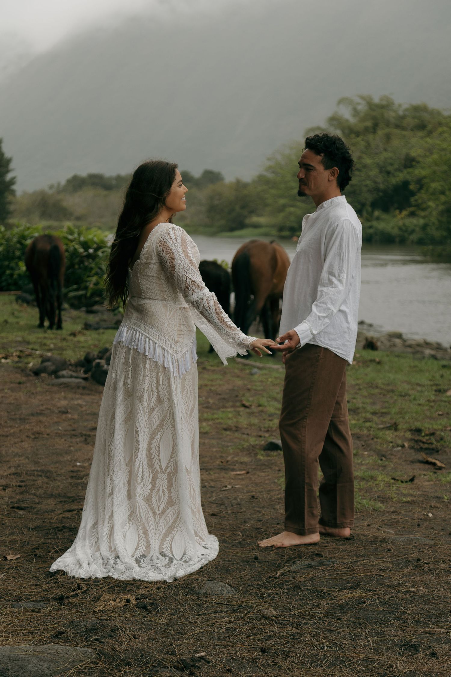 Couple in white lace dress and white shirt hold hands near horses by a misty river.