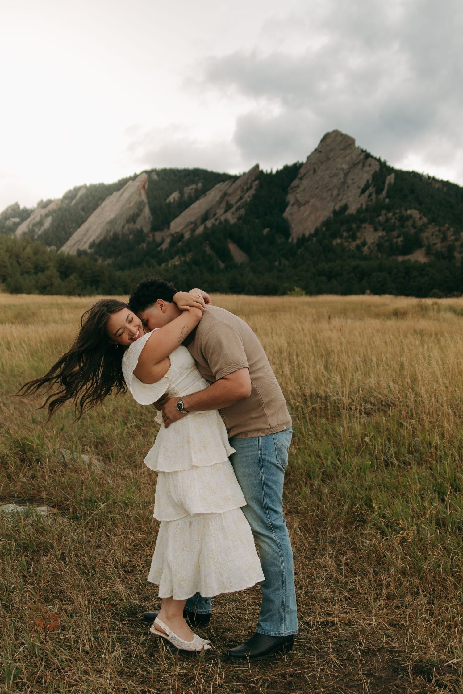 Man embracing woman in white tiered dress in open field with mountain backdrop at dusk.
