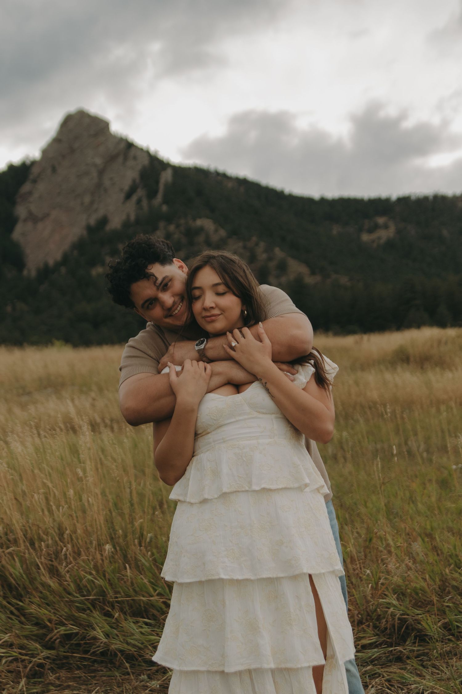 Couple embracing in golden meadow with rocky mountain backdrop, woman in white ruffled dress, romantic outdoor portrait.