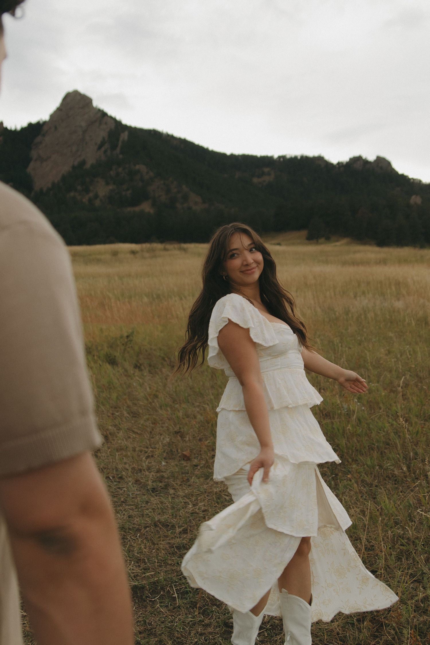 Woman in white tiered dress twirling in mountain meadow field with rocky flatirons backdrop.