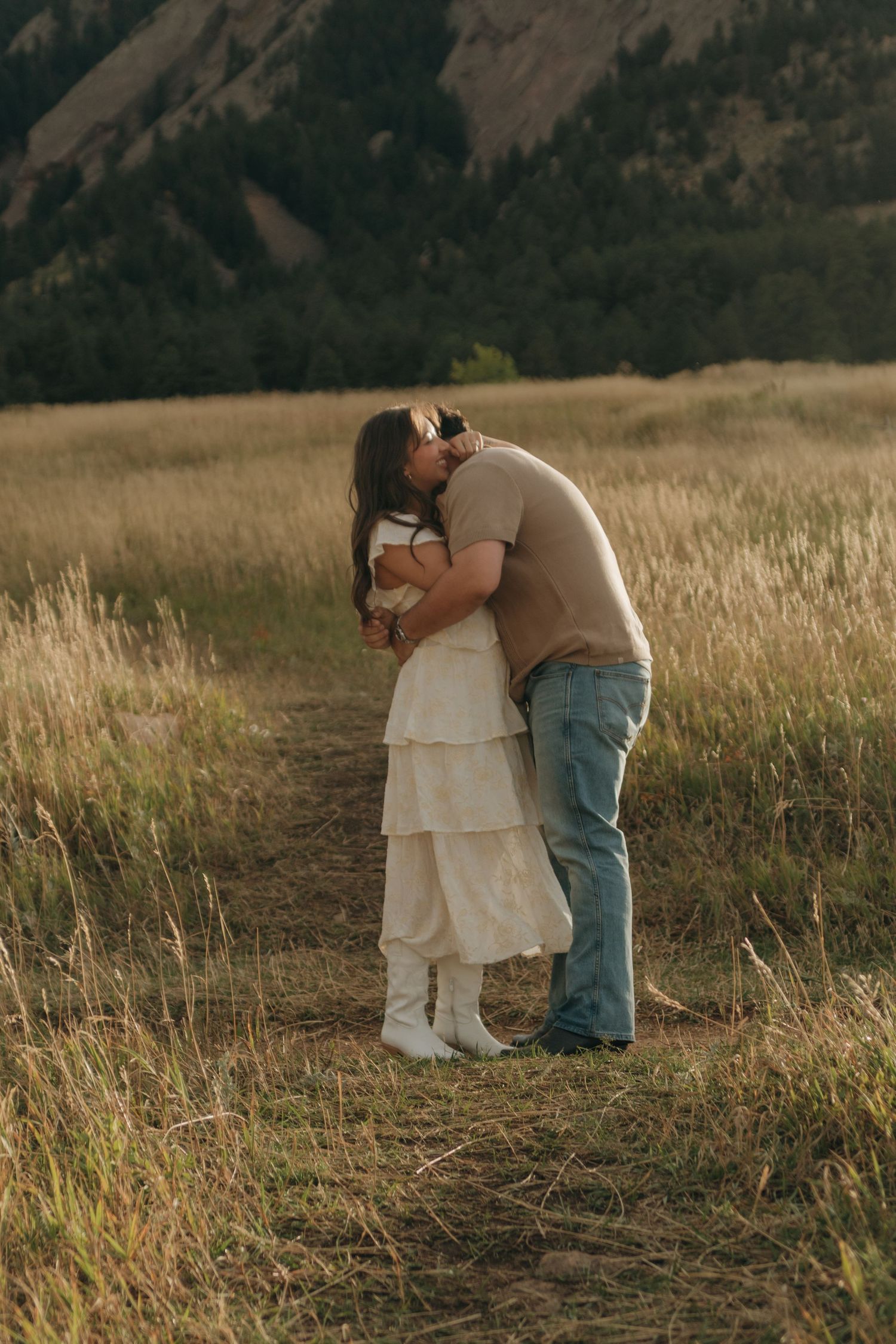 Couple sharing a romantic kiss in a golden meadow with mountain backdrop during sunset golden hour.