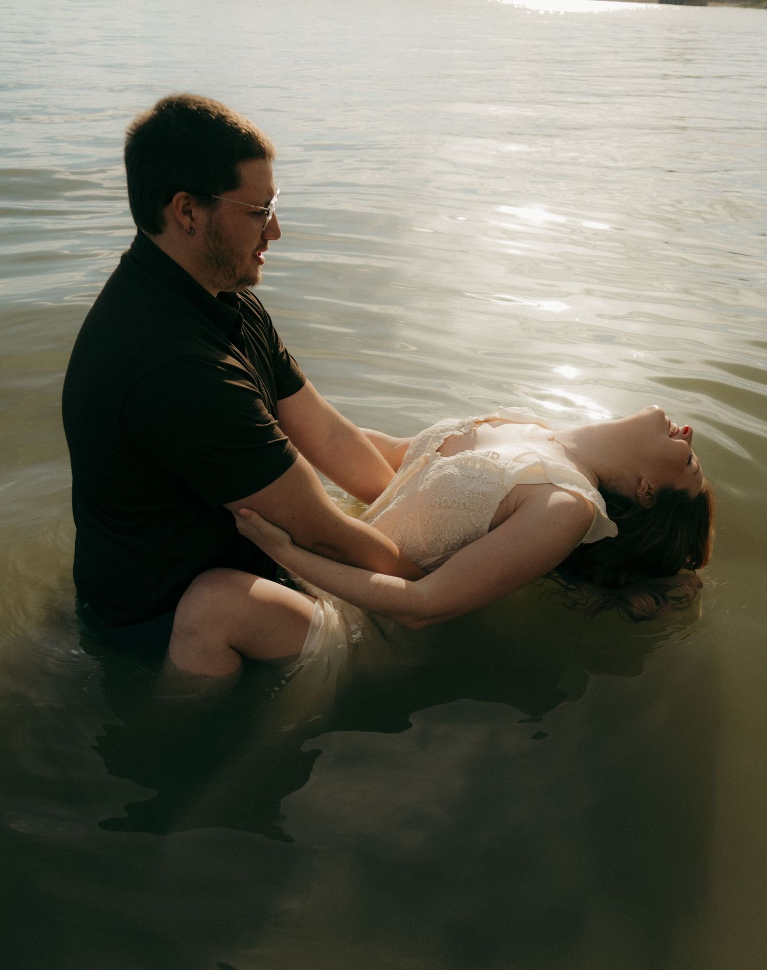 Man gently holding a woman as she floats on her back in a shimmering sunlit lake.