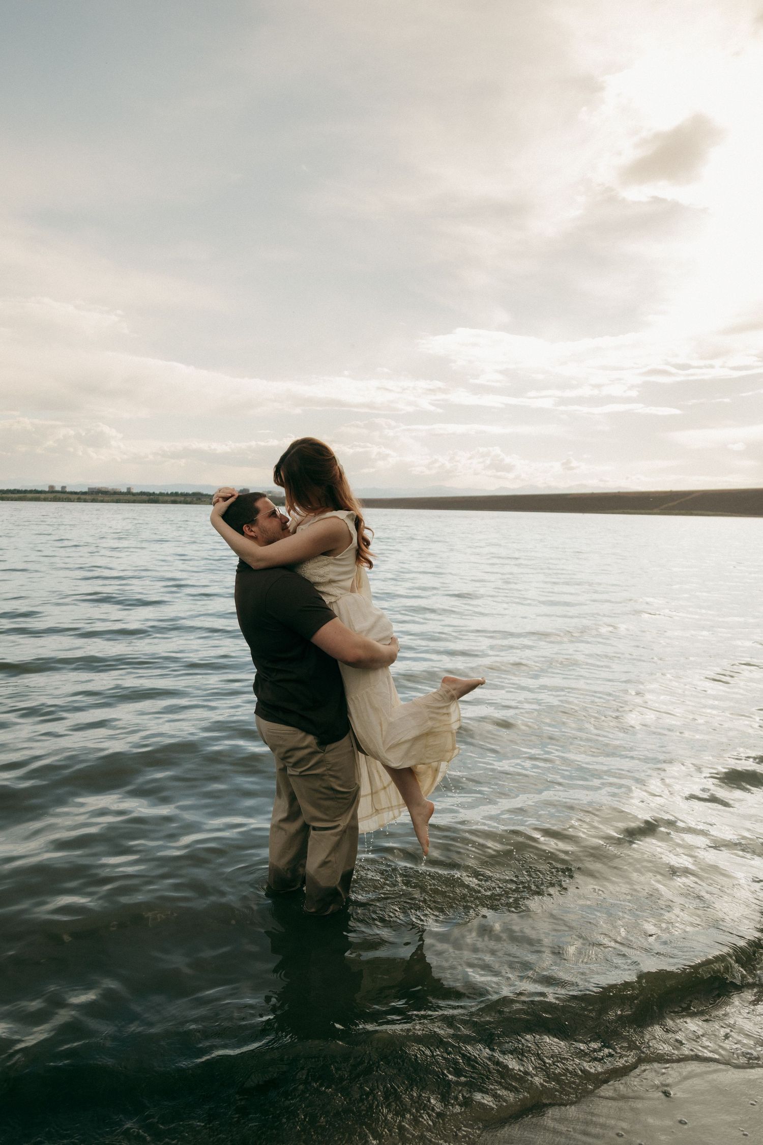 Couple embracing romantically in shallow lake water at golden sunset, man lifting woman in white dress.