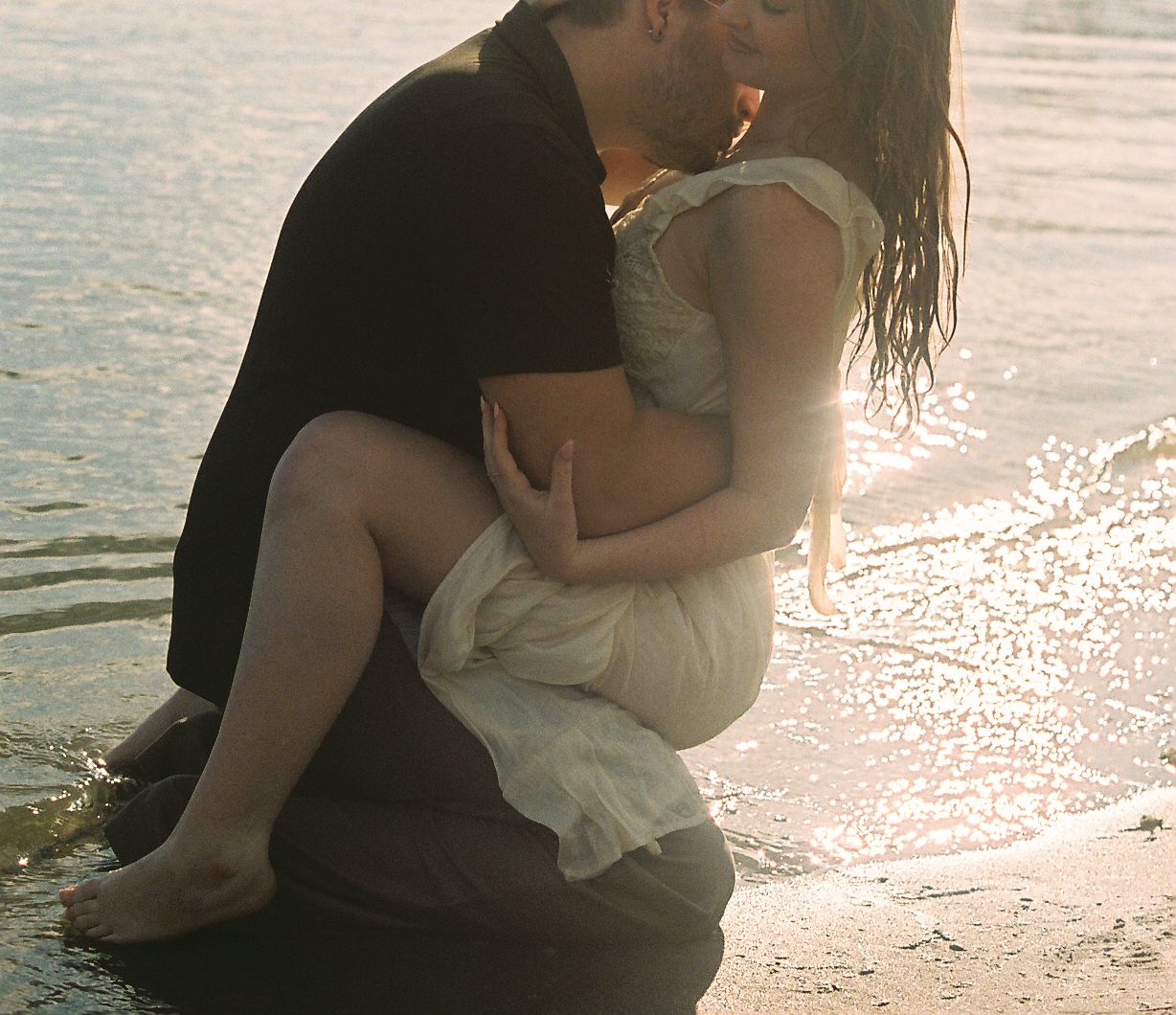 Man cradling woman in white dress near shoreline, sharing a kiss in golden sunset light.