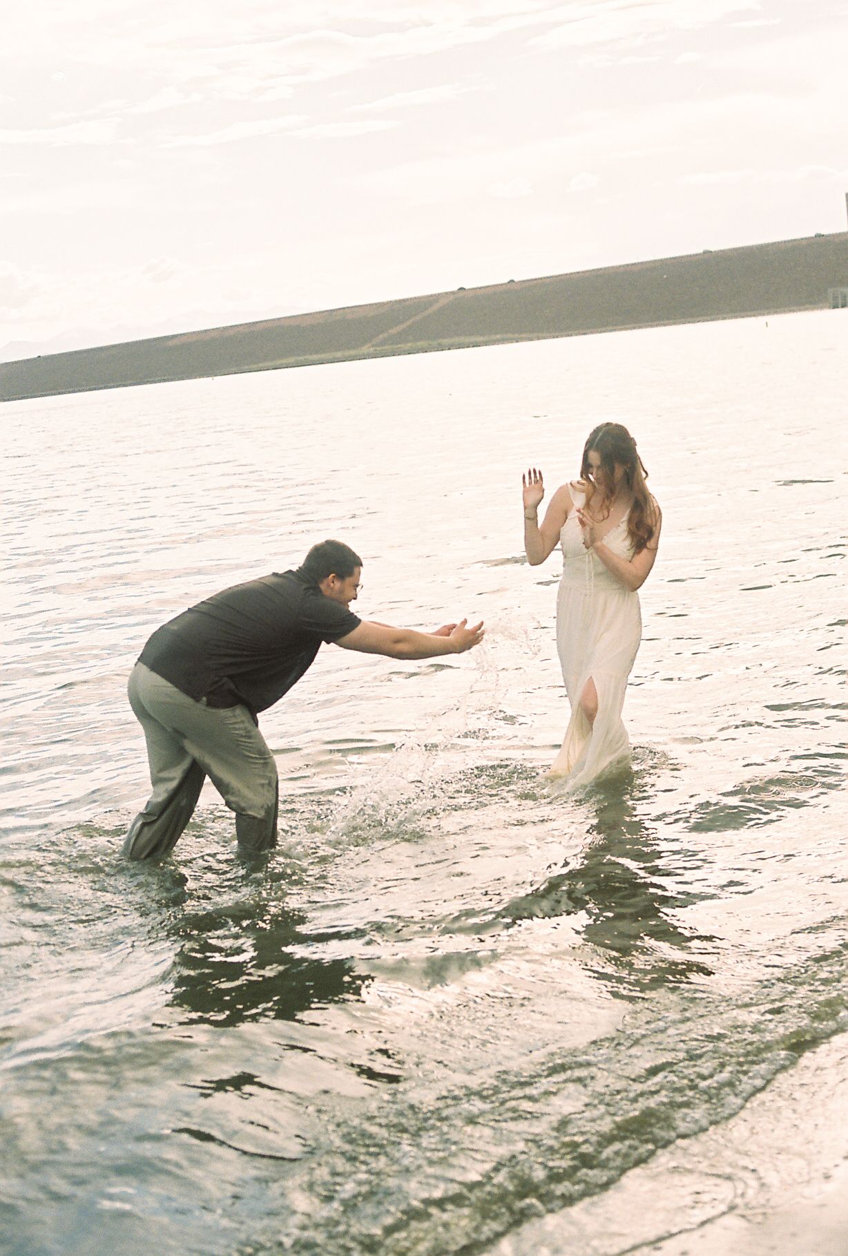 Playful couple splashing water at each other while wading in a serene lake.
