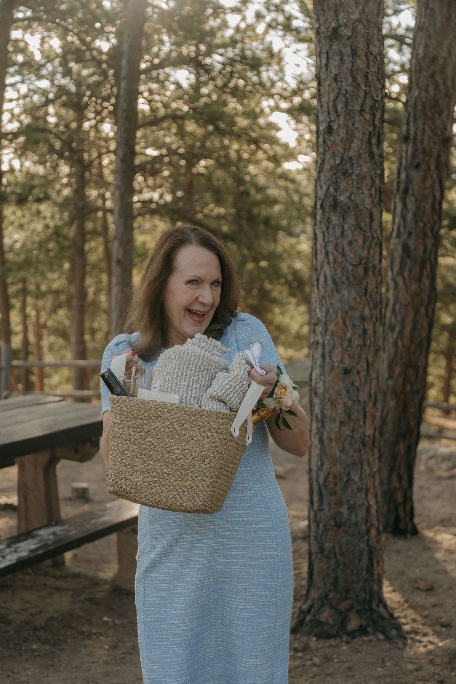 Smiling woman in blue dress holds woven basket filled with wedding supplies in a pine forest.