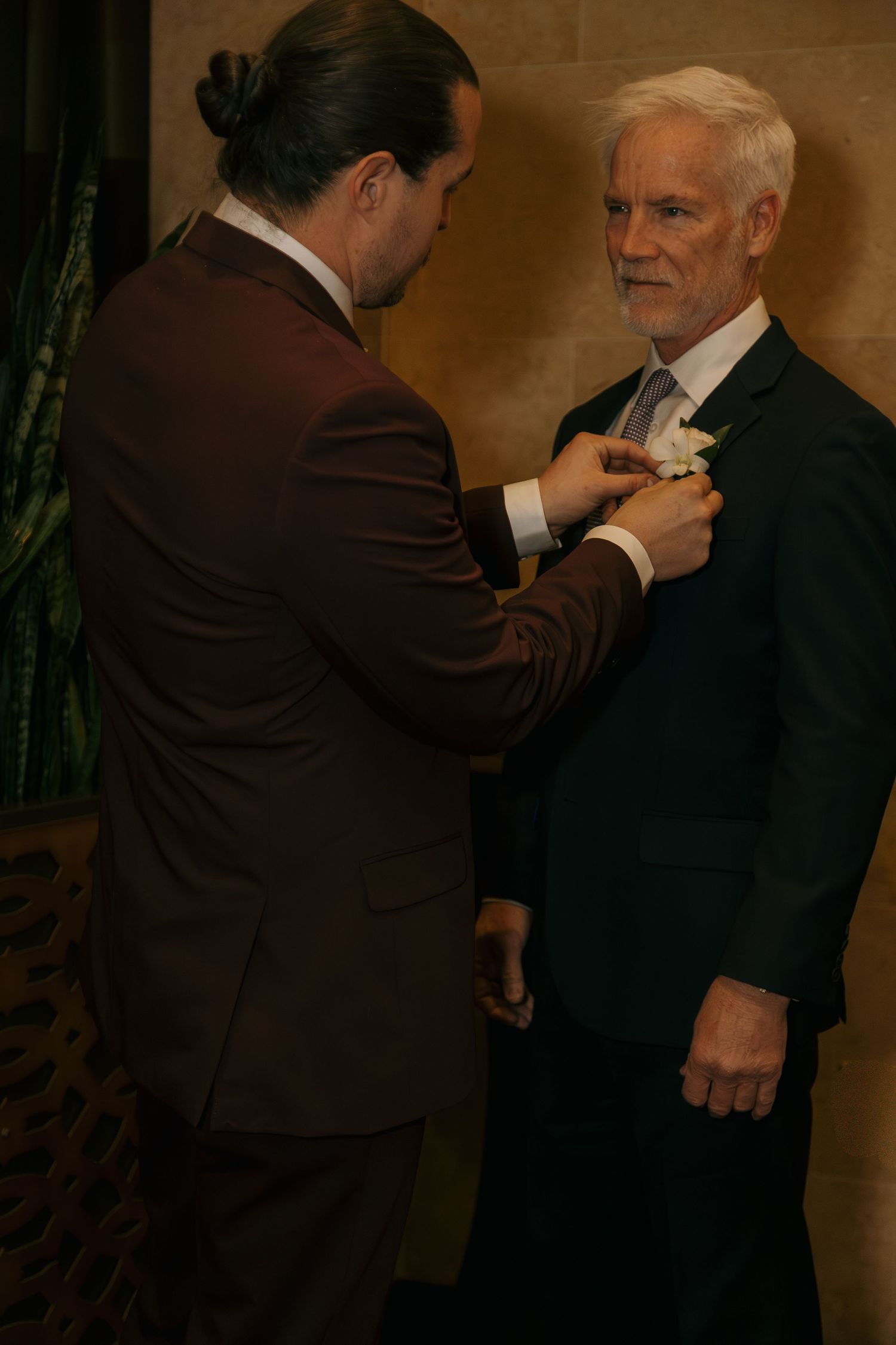 Man in burgundy suit pins boutonniere on older gentleman in navy suit at formal wedding event.