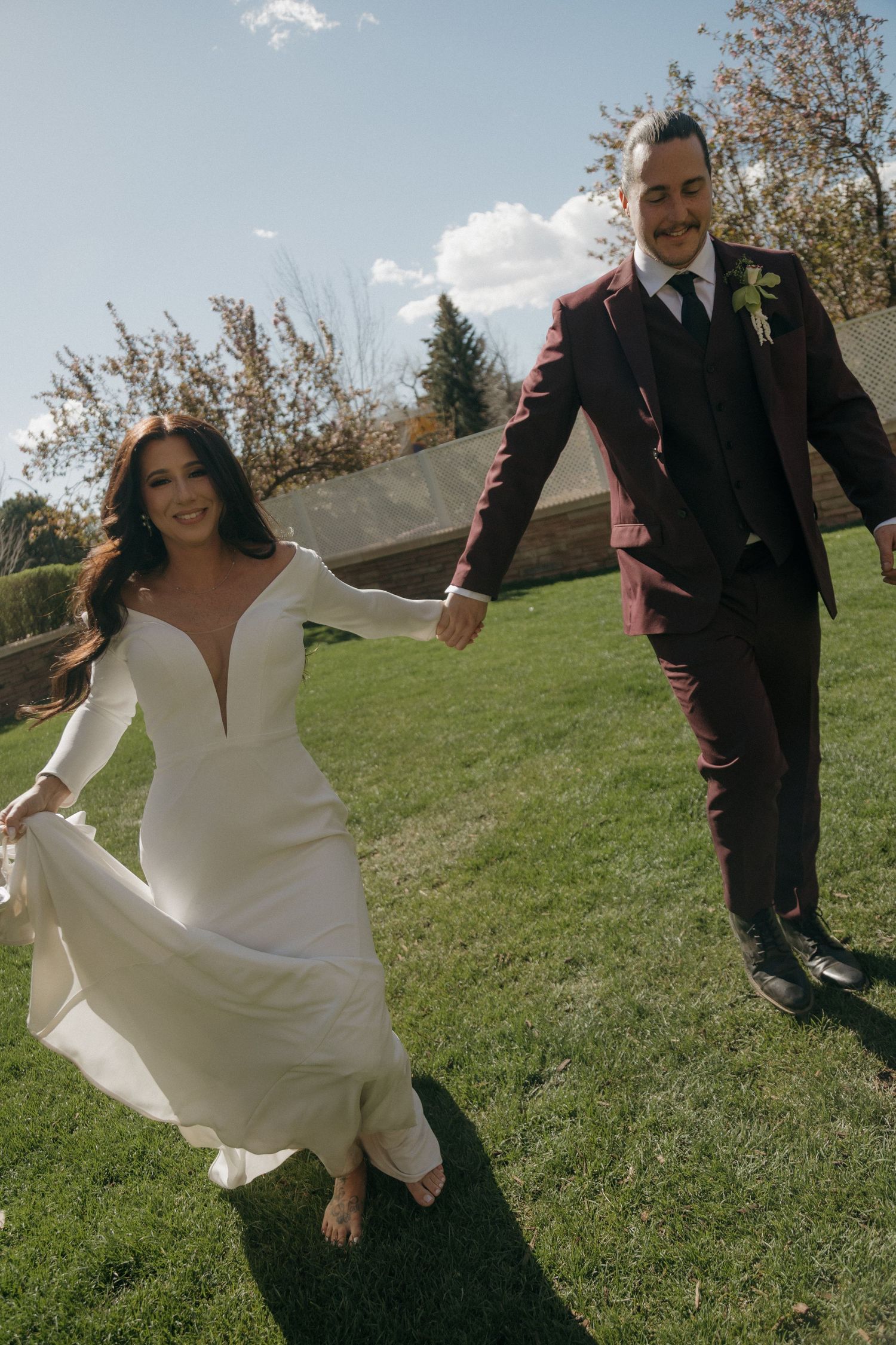 Smiling bride in white gown and groom in burgundy suit holding hands running across green lawn.