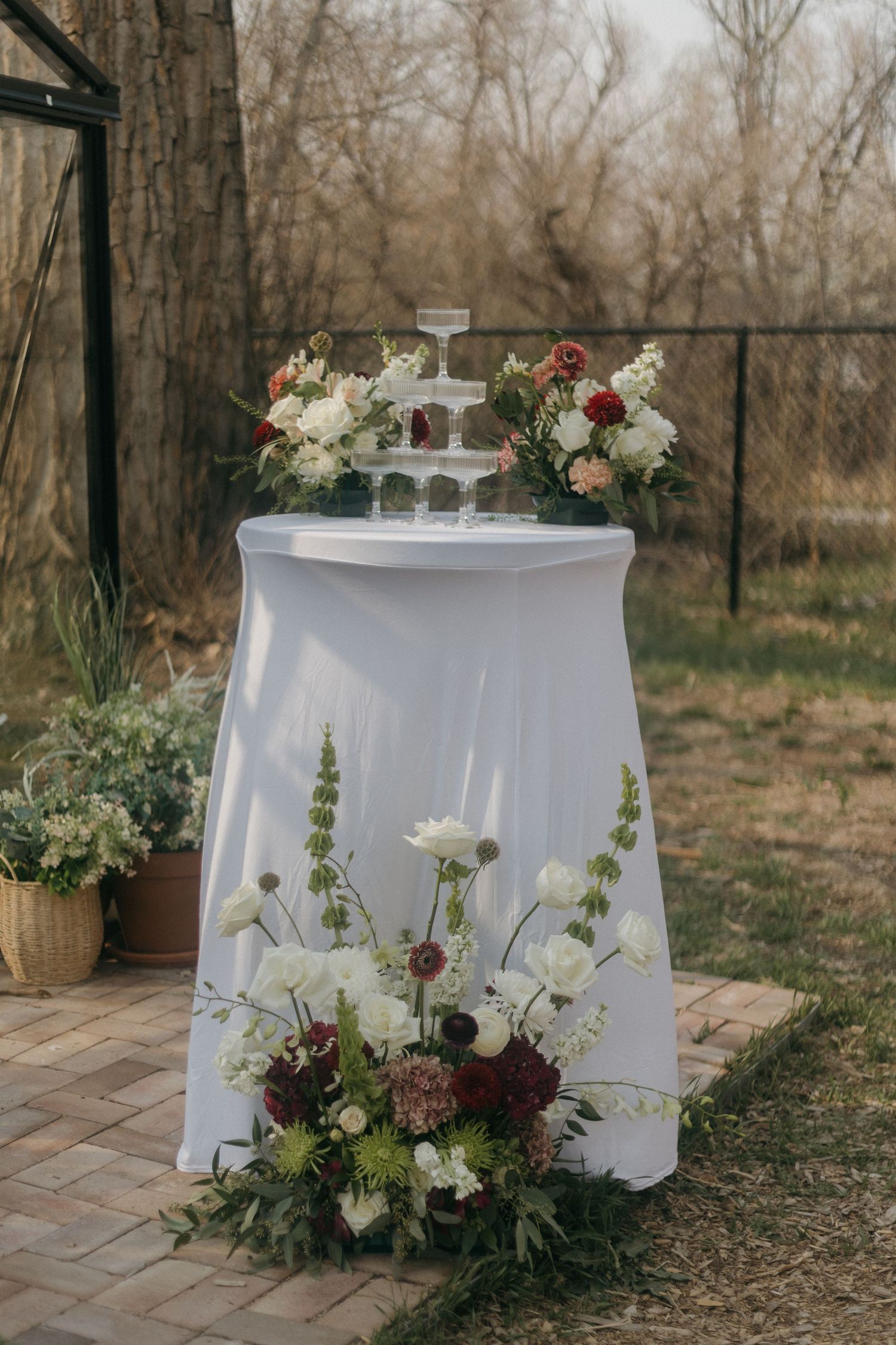 Elegant champagne tower centerpiece surrounded by white and burgundy floral arrangements on a draped table.