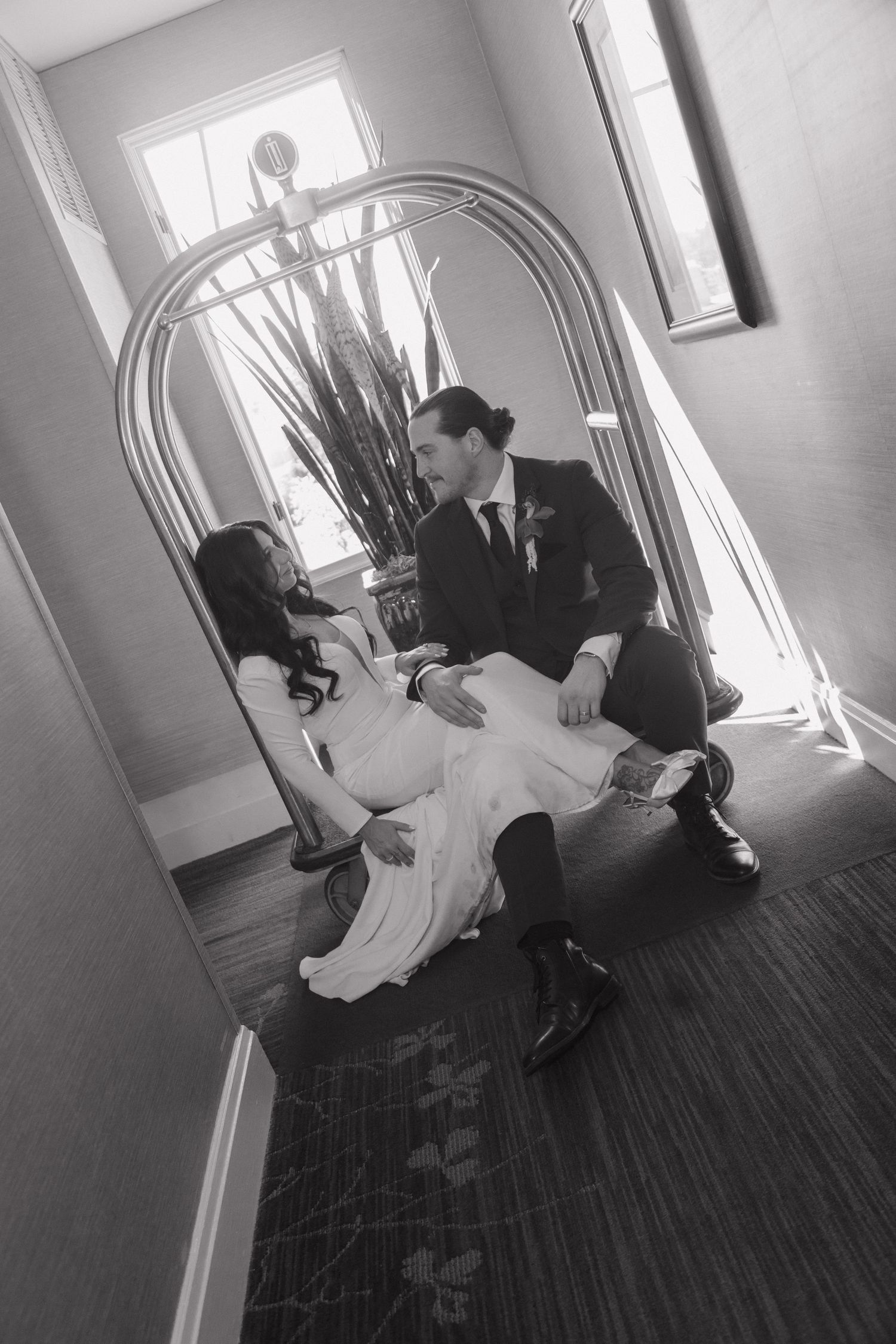 Black and white photo of bride and groom sitting together on a hotel floor by a luggage cart.