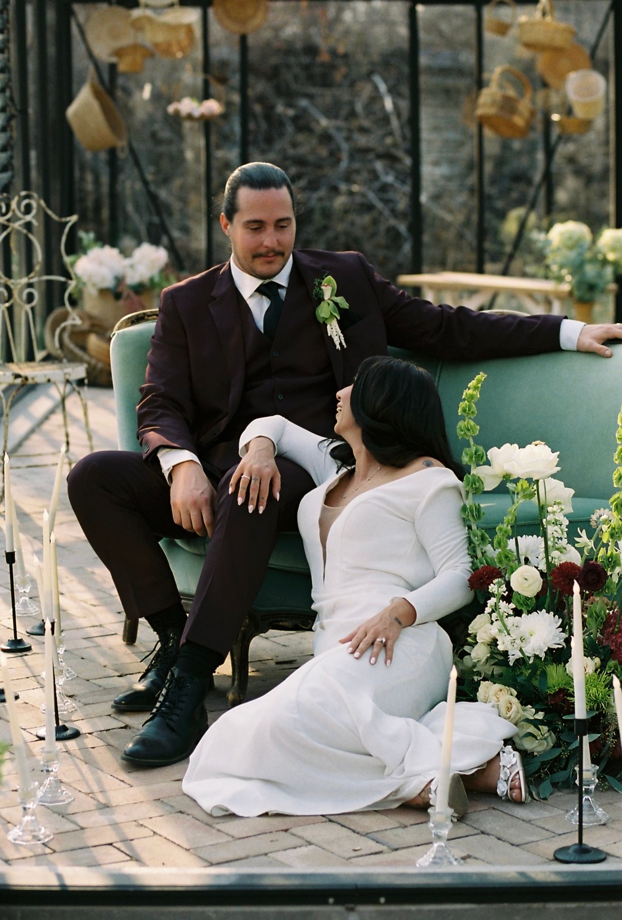 Bride and groom relax on a teal vintage sofa inside a glass greenhouse surrounded by lush floral arrangements.
