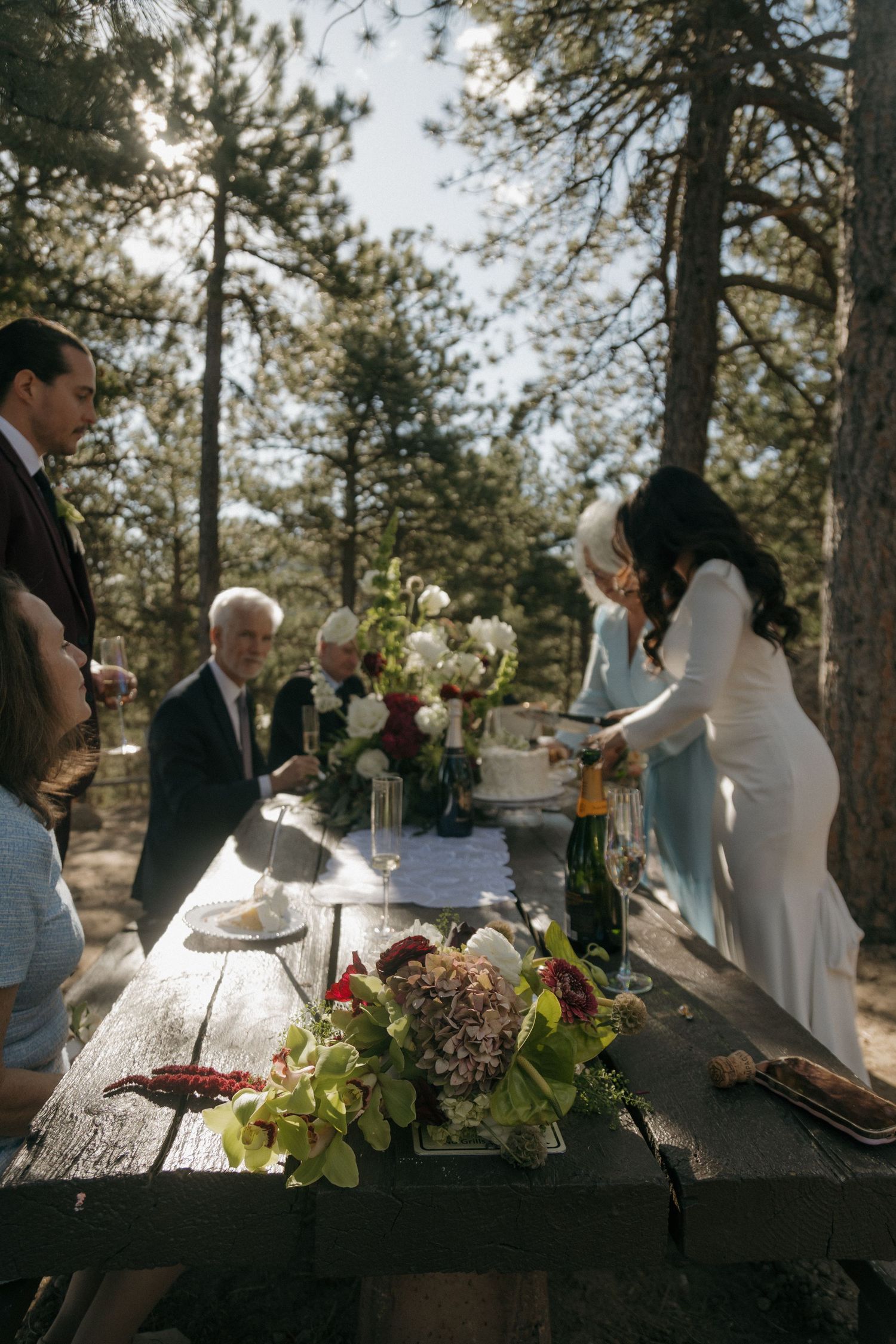 Wedding guests gather around a rustic wooden table adorned with flowers and champagne in a sunlit forest setting.