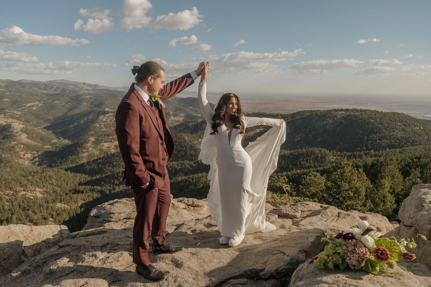 Groom in burgundy suit twirling bride in white dress on a rocky mountain overlook with scenic valley views.