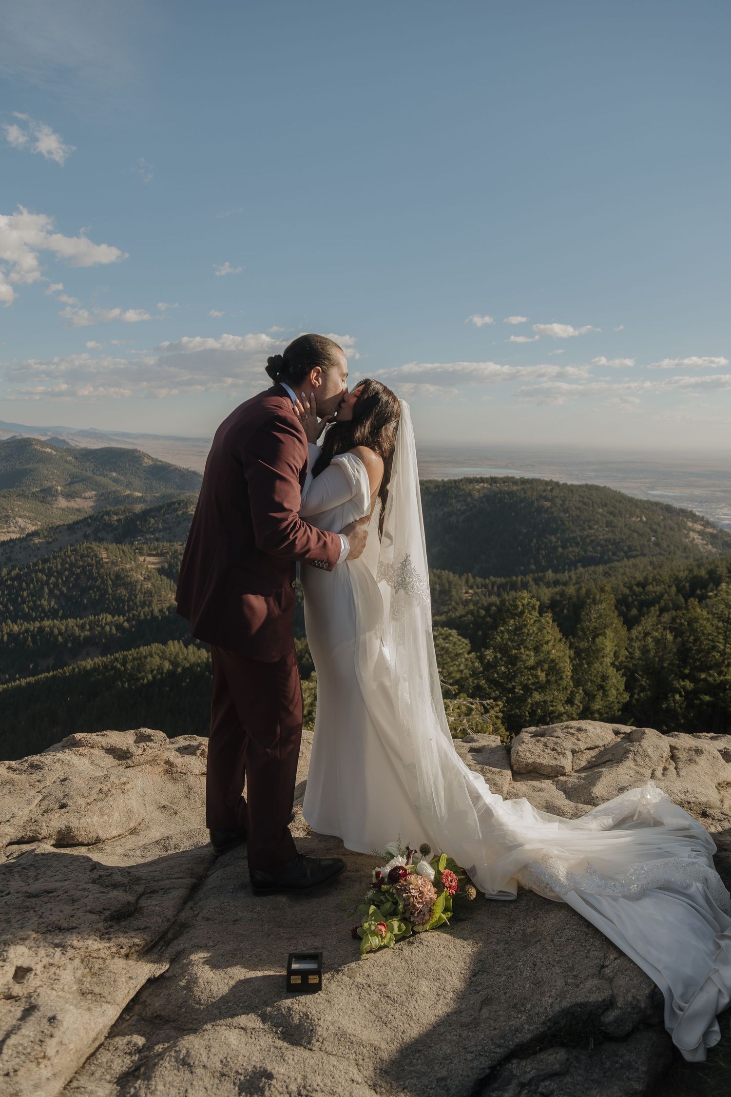 Bride and groom share first kiss on rocky mountain summit with ring box and floral bouquet nearby.
