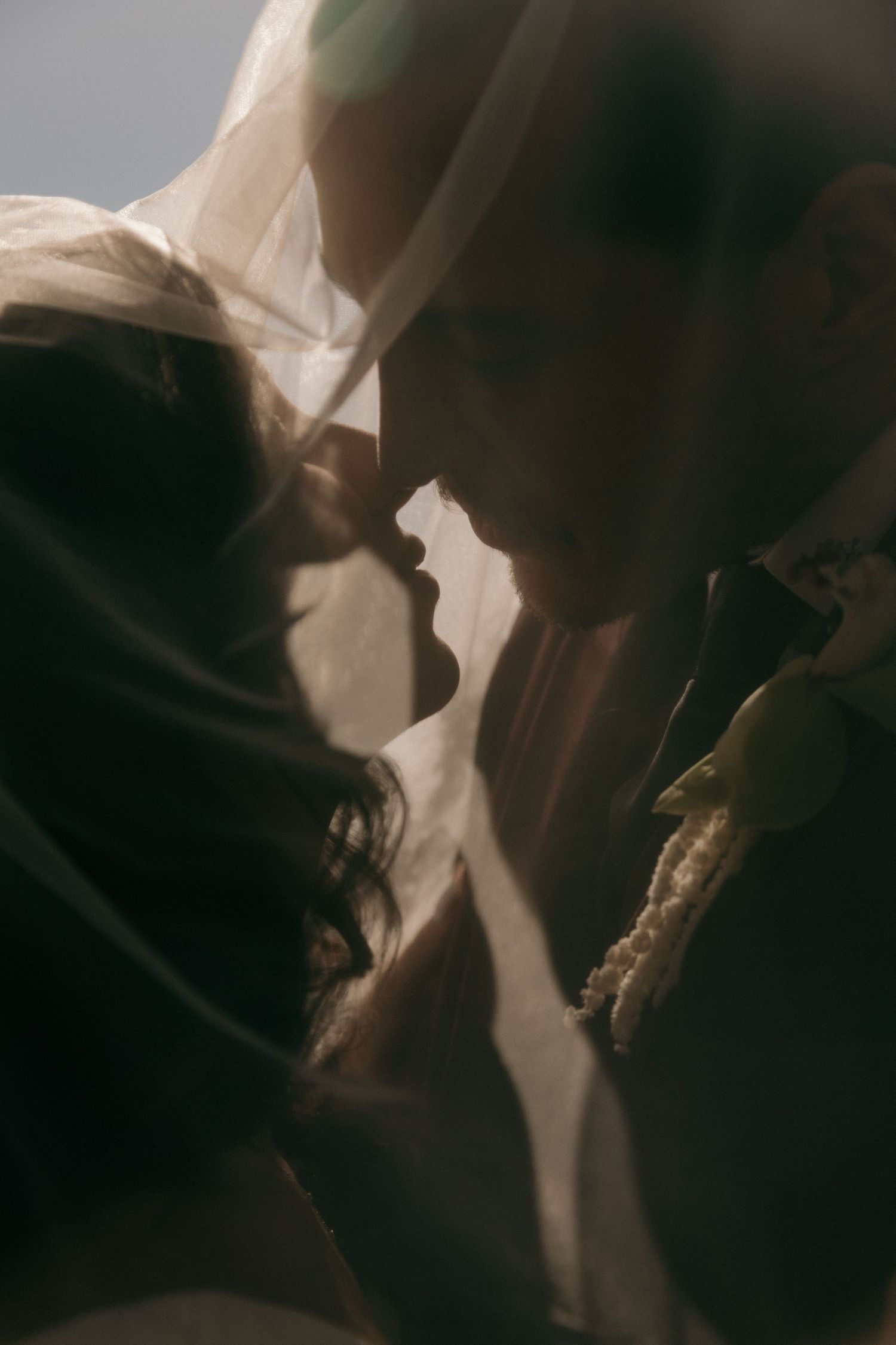 Intimate close-up of bride and groom nearly kissing under sheer wedding veil in soft dramatic lighting.