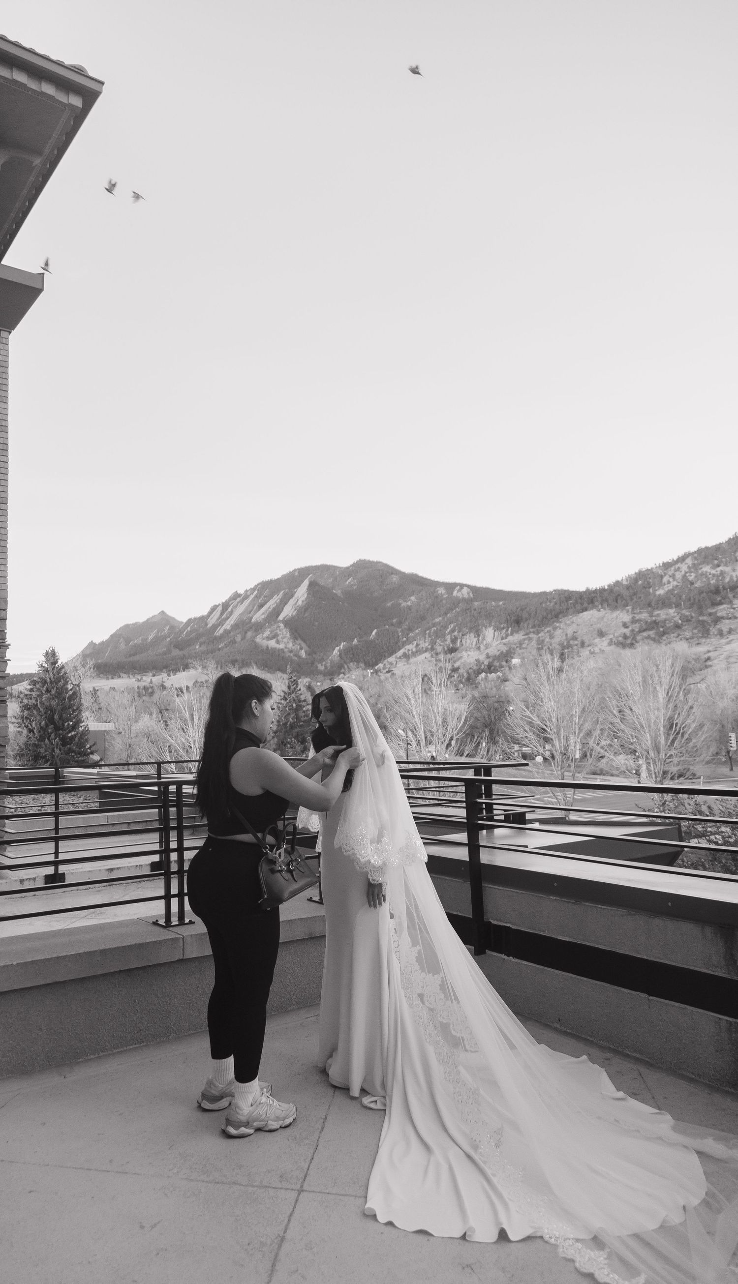 Bride in white gown and veil being assisted on balcony with mountain backdrop in black and white.