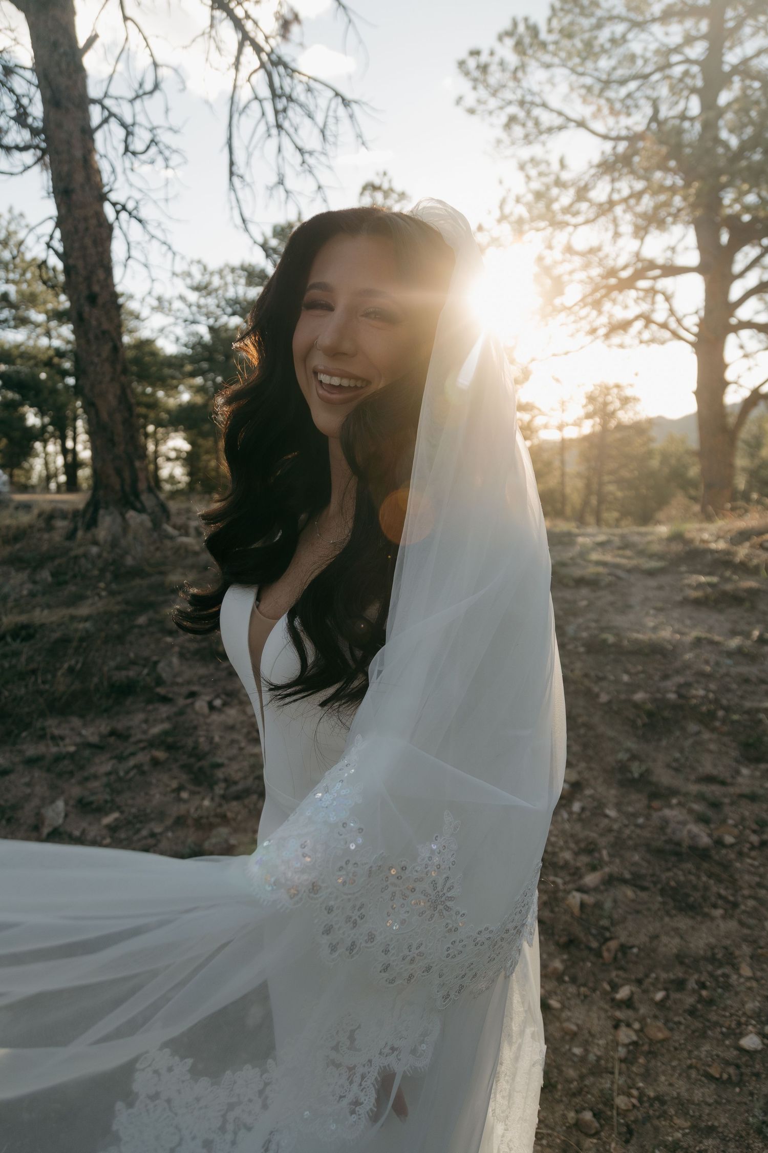 Smiling bride in white dress and embroidered veil posing in pine forest with golden sunset backlight.