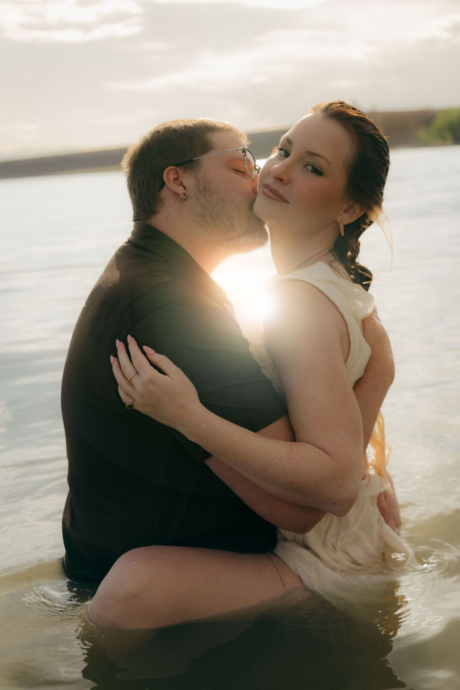 Couple embracing romantically in water at sunset, man kissing woman's cheek, both in formal attire with golden backlight.