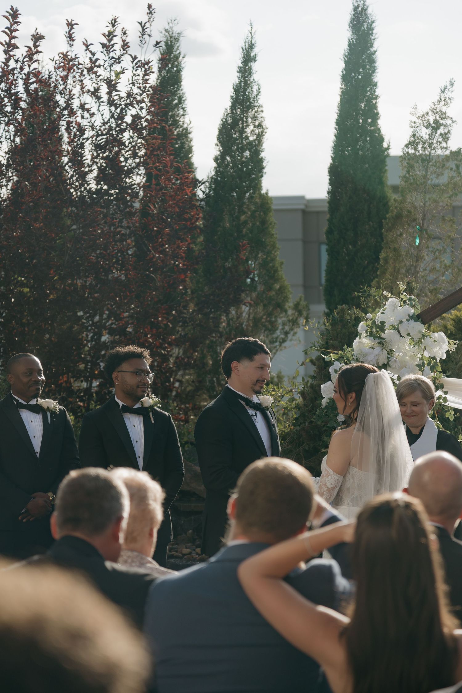 Groom smiles at bride during outdoor wedding ceremony with groomsmen and cypress trees in background.