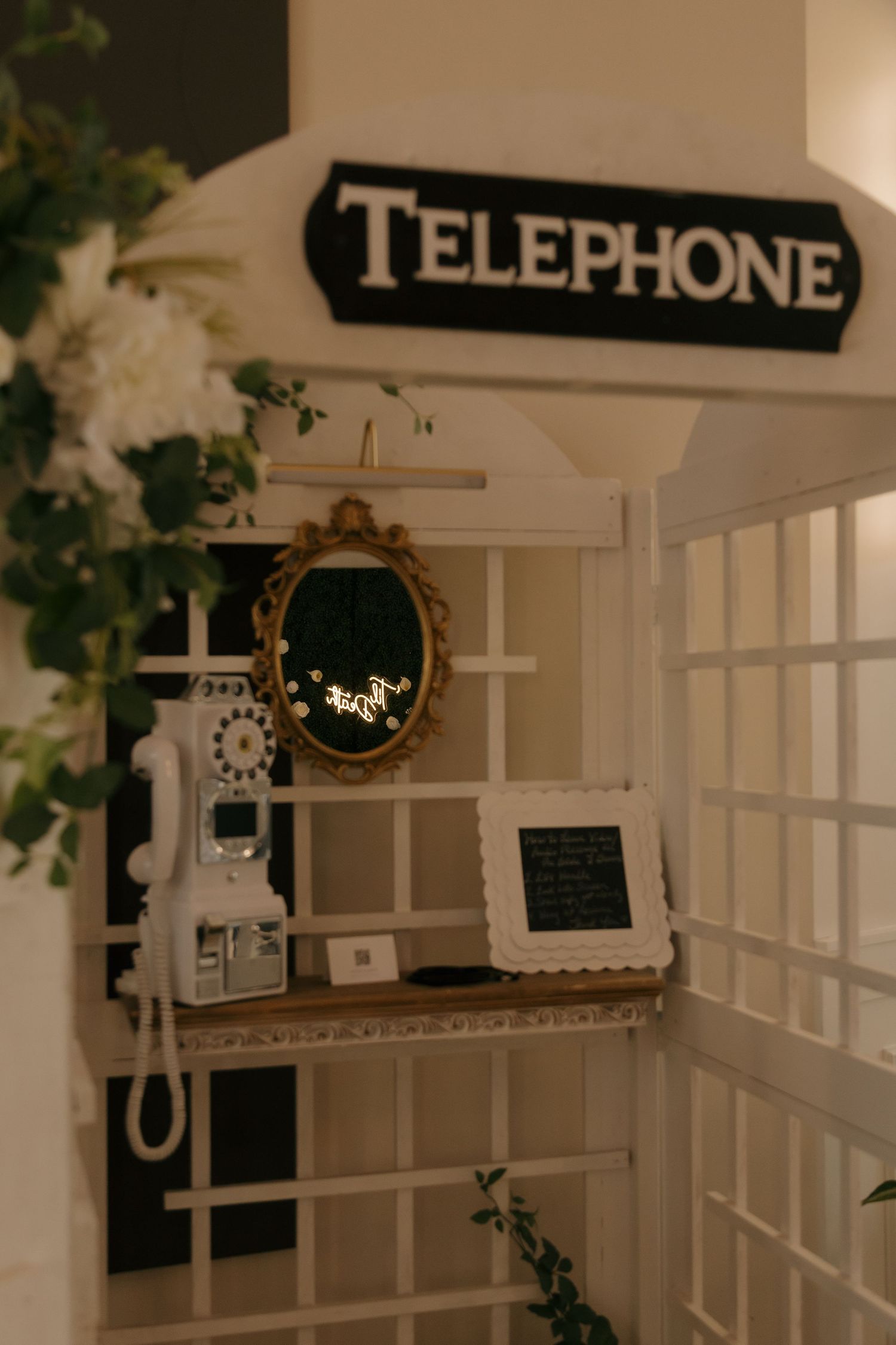Vintage white telephone booth decorated with flowers, ornate mirror, and rotary phone for elegant event decor.