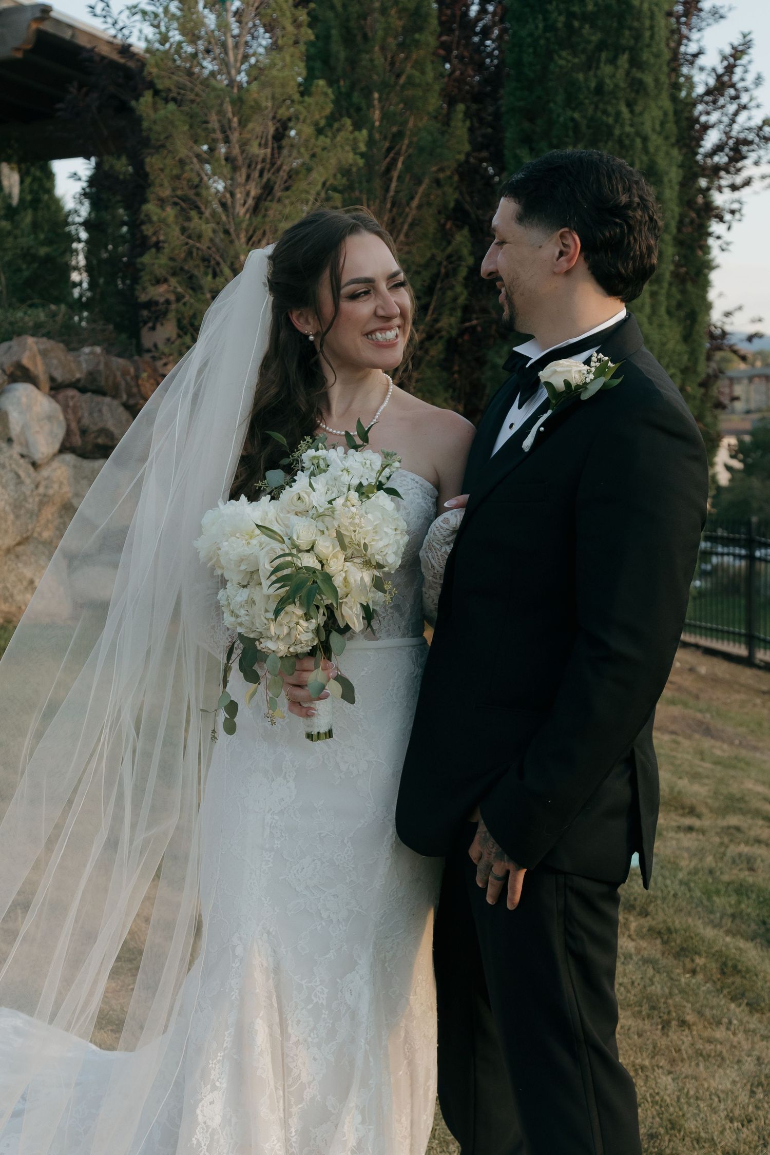 Smiling bride in white gown with veil and bouquet stands with groom in black tuxedo outdoors.
