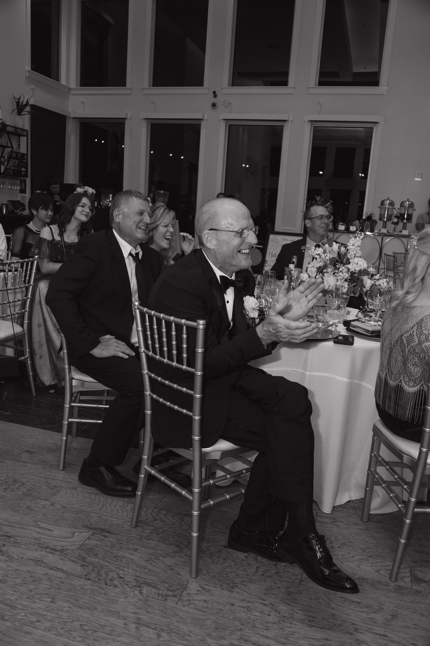 Black and white photo of wedding guests laughing and clapping during reception speeches at elegant venue.