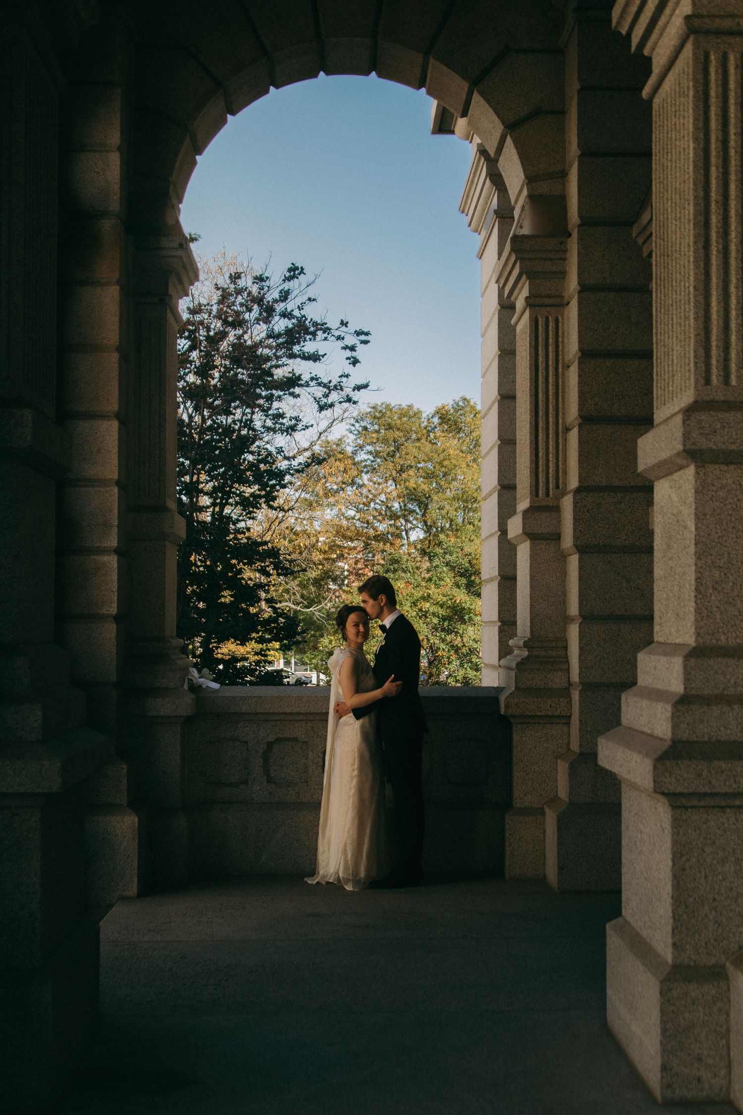A romantic moment captured through an arched colonnade with trees visible in the background.