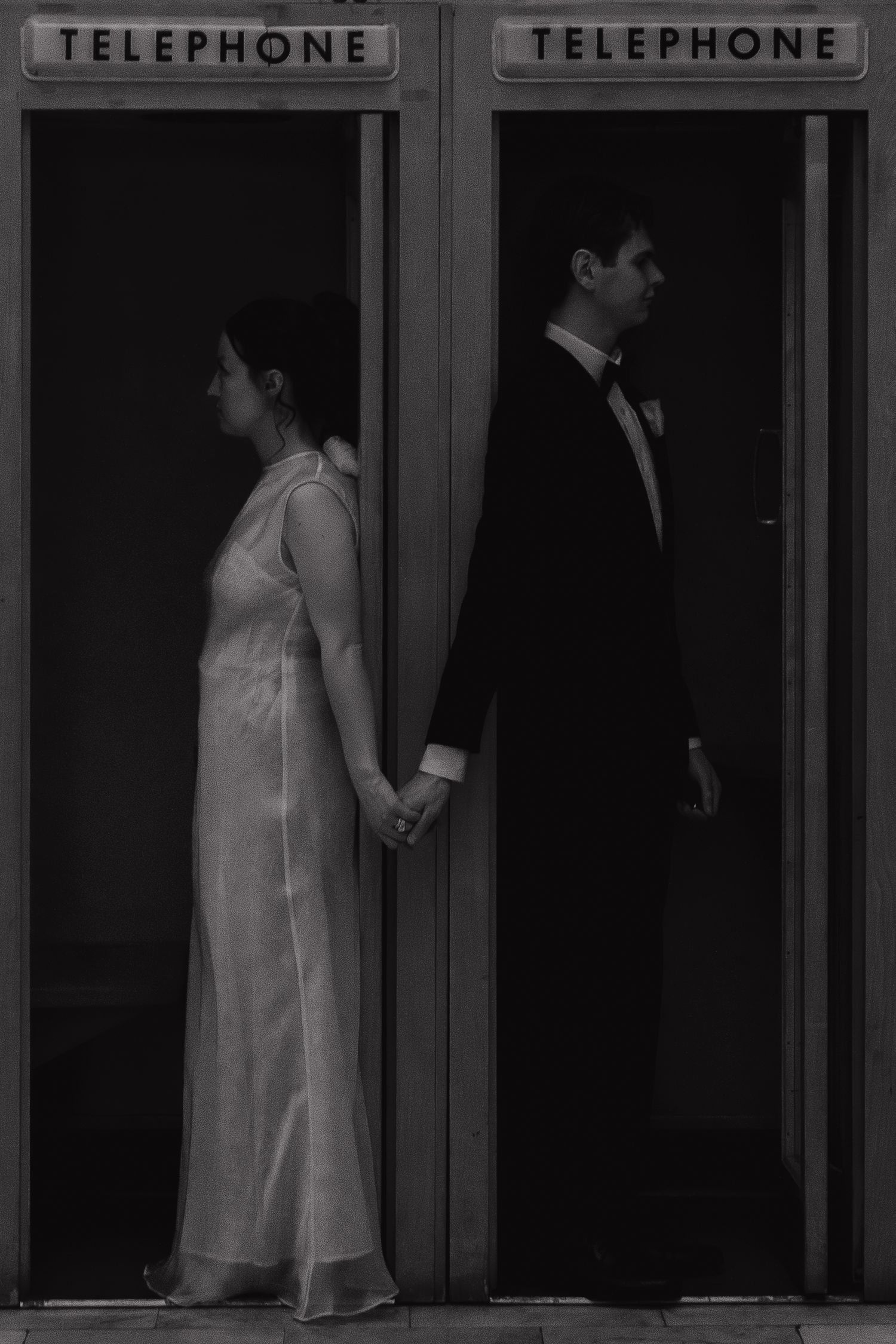 A couple stands back to back in adjacent phone booths, holding hands through the divider in a black and white photo.