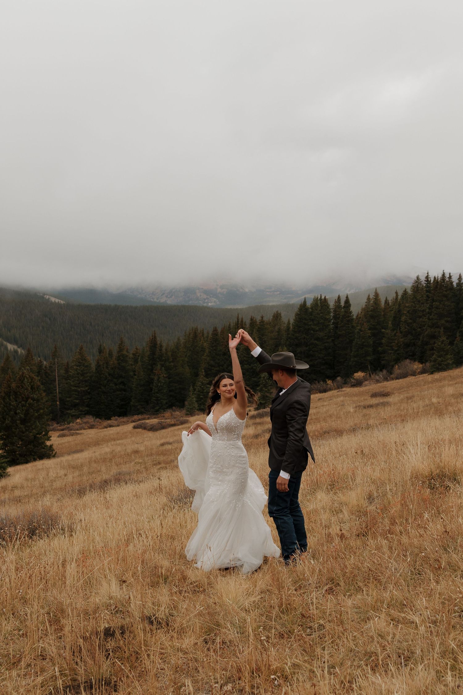 A couple dances together in a mountain meadow with evergreen trees and cloudy skies.