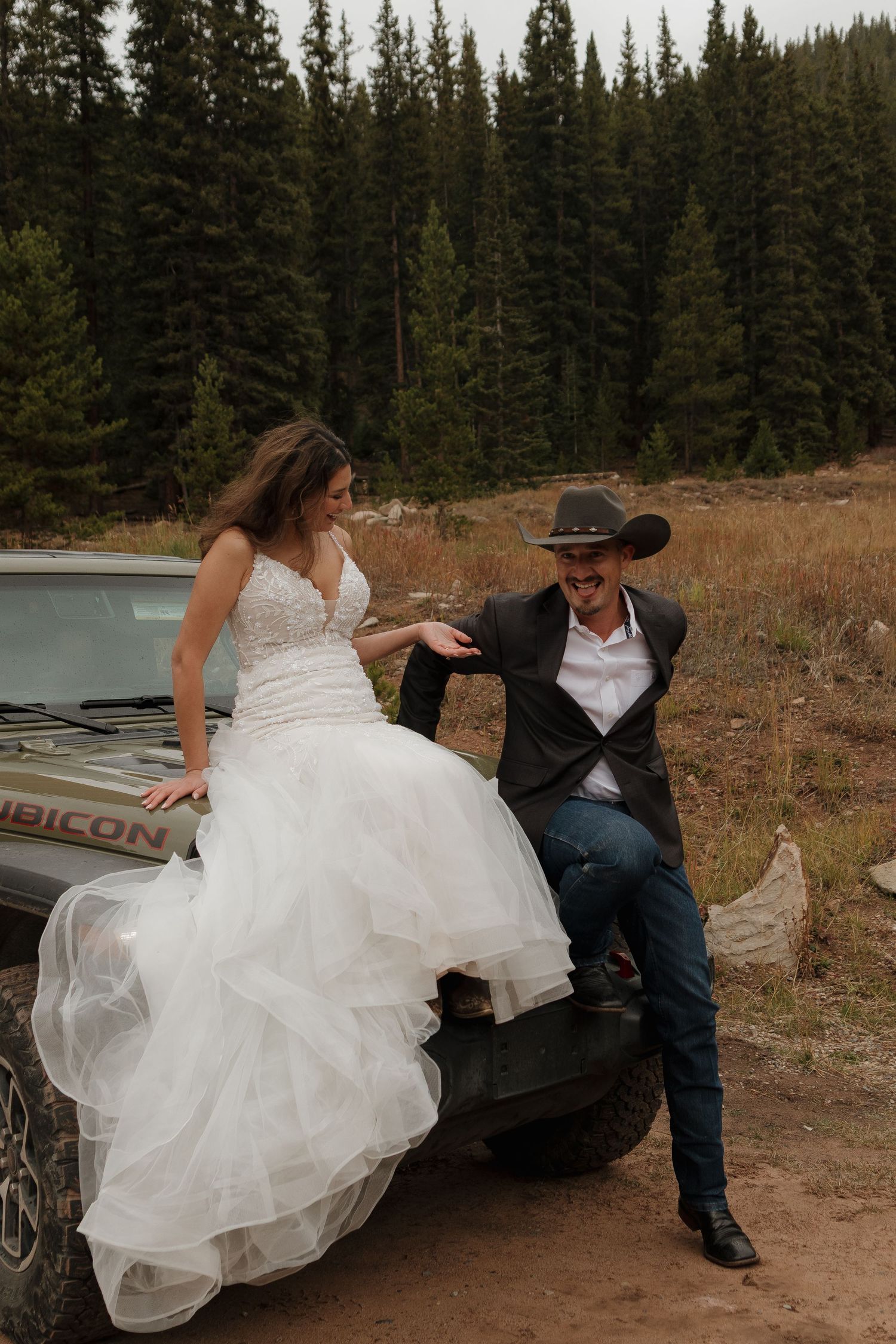 A couple in wedding attire sits casually on a rustic bench surrounded by pine trees.
