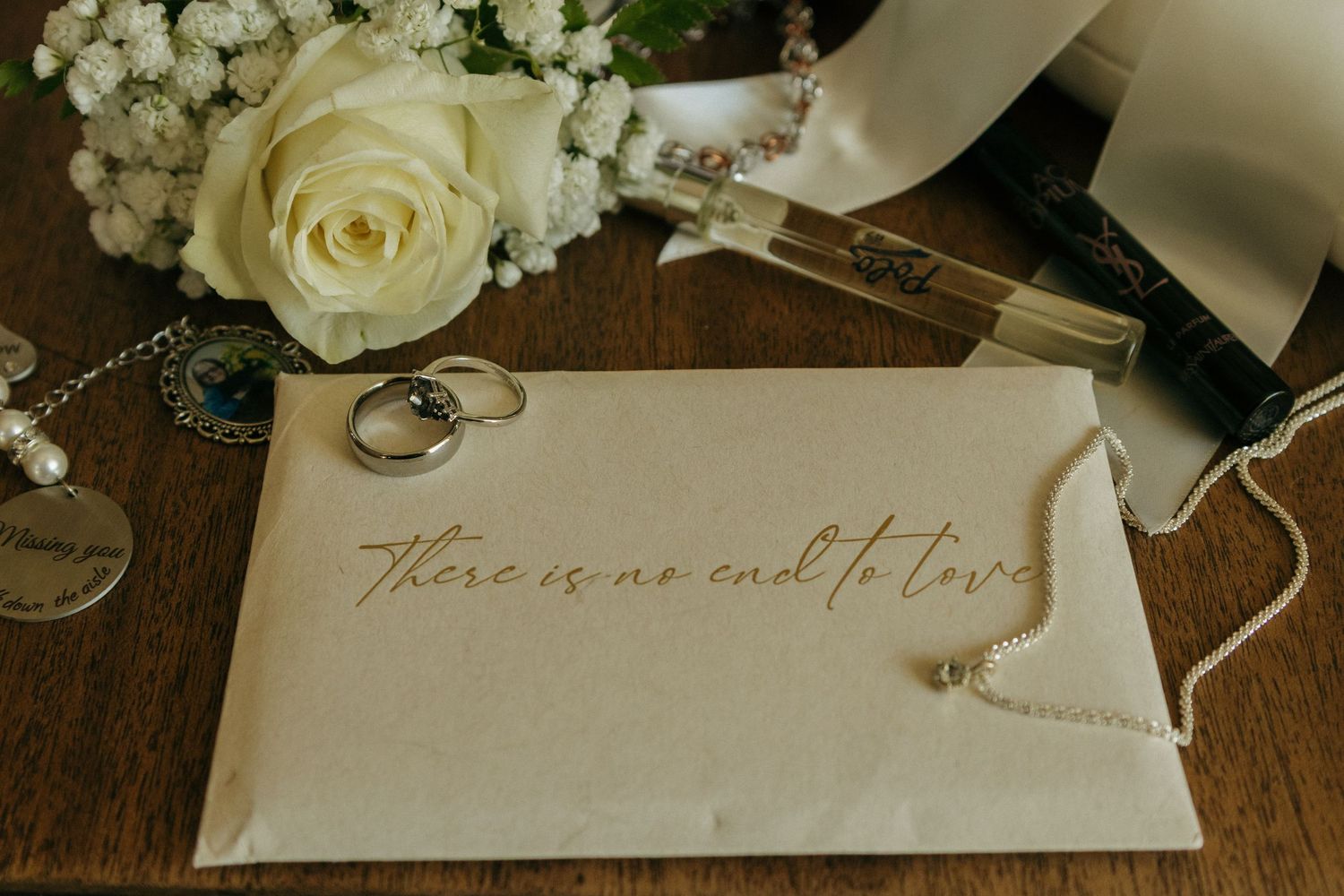 Wedding rings and white rose bouquet next to a handwritten note that reads 'There is no end to love' on wooden surface.